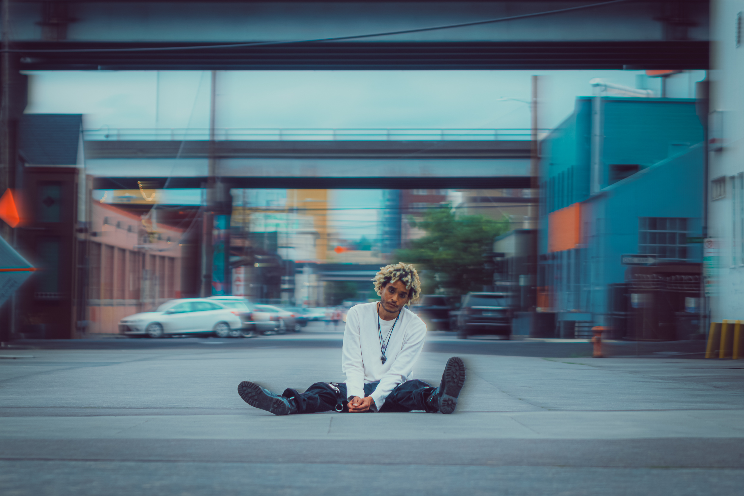 A young artist sitting cross-legged on a city street, wearing a white long-sleeve shirt and black pants, with buildings and vehicles in the background.