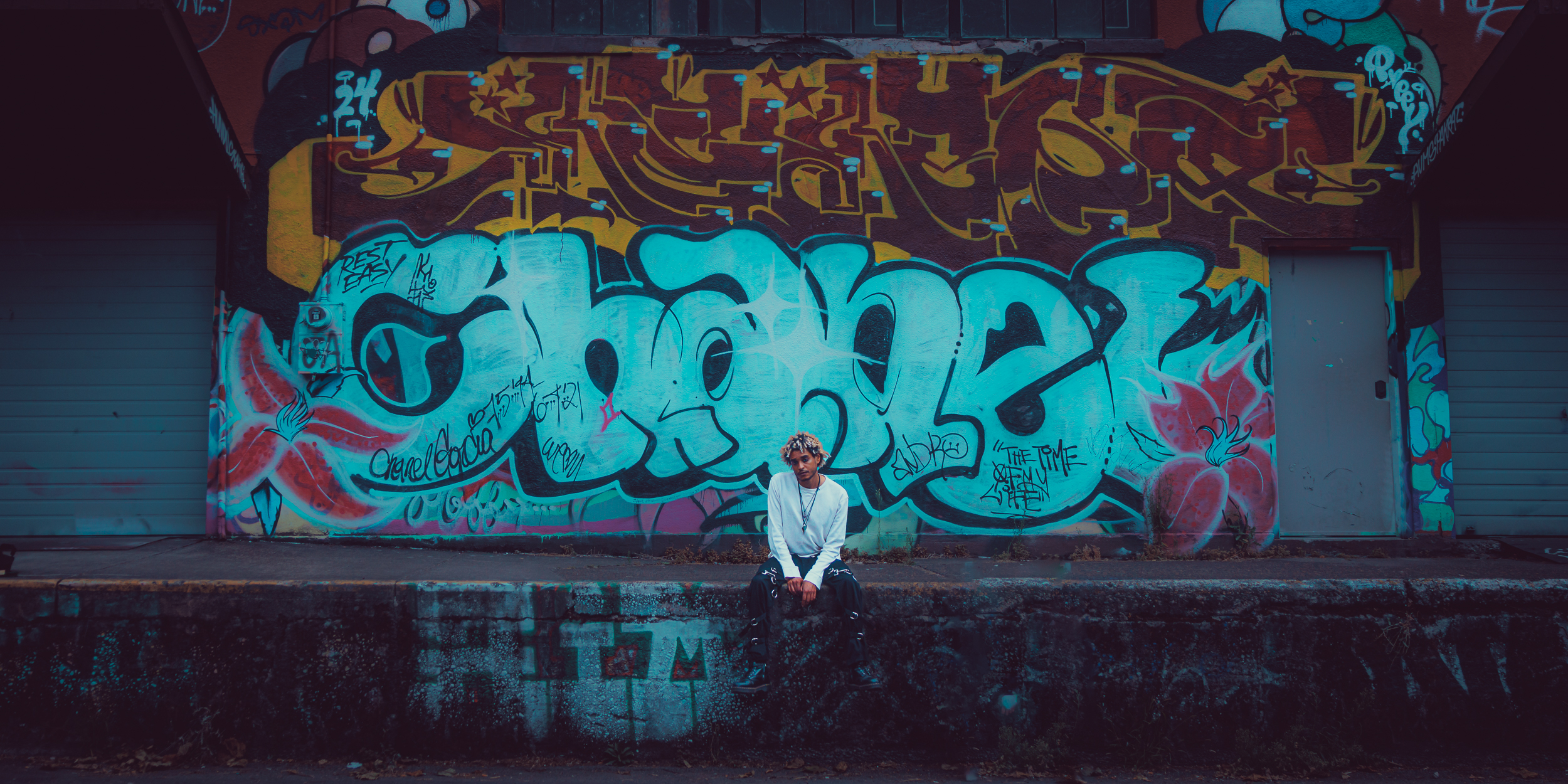 A young artist sitting on a ledge in front of a vibrant graffiti wall featuring the word 'Chanel' and various colorful designs.