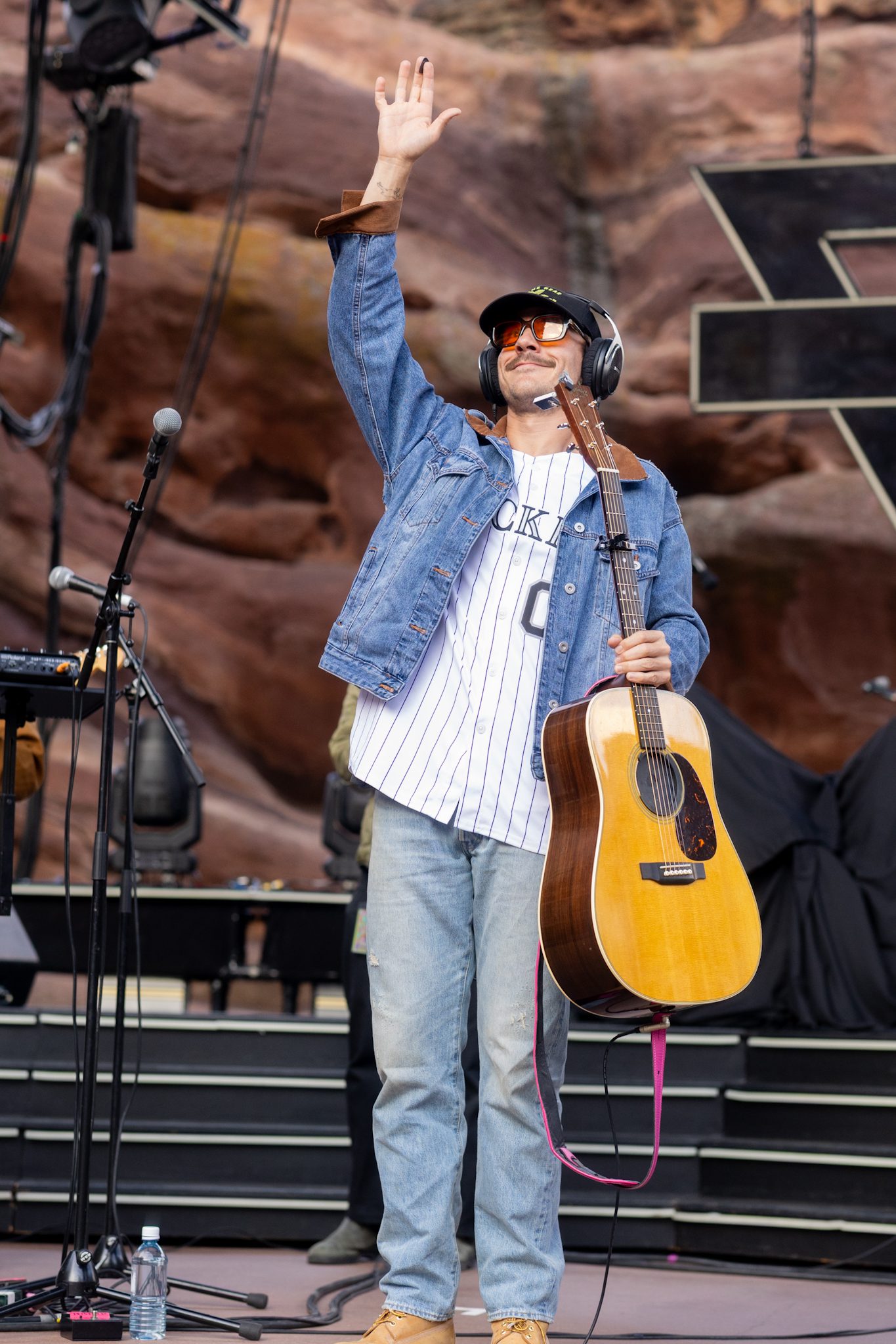 A musician on stage at Red Rocks, wearing a denim jacket and sunglasses, waving to the crowd while holding an acoustic guitar.
