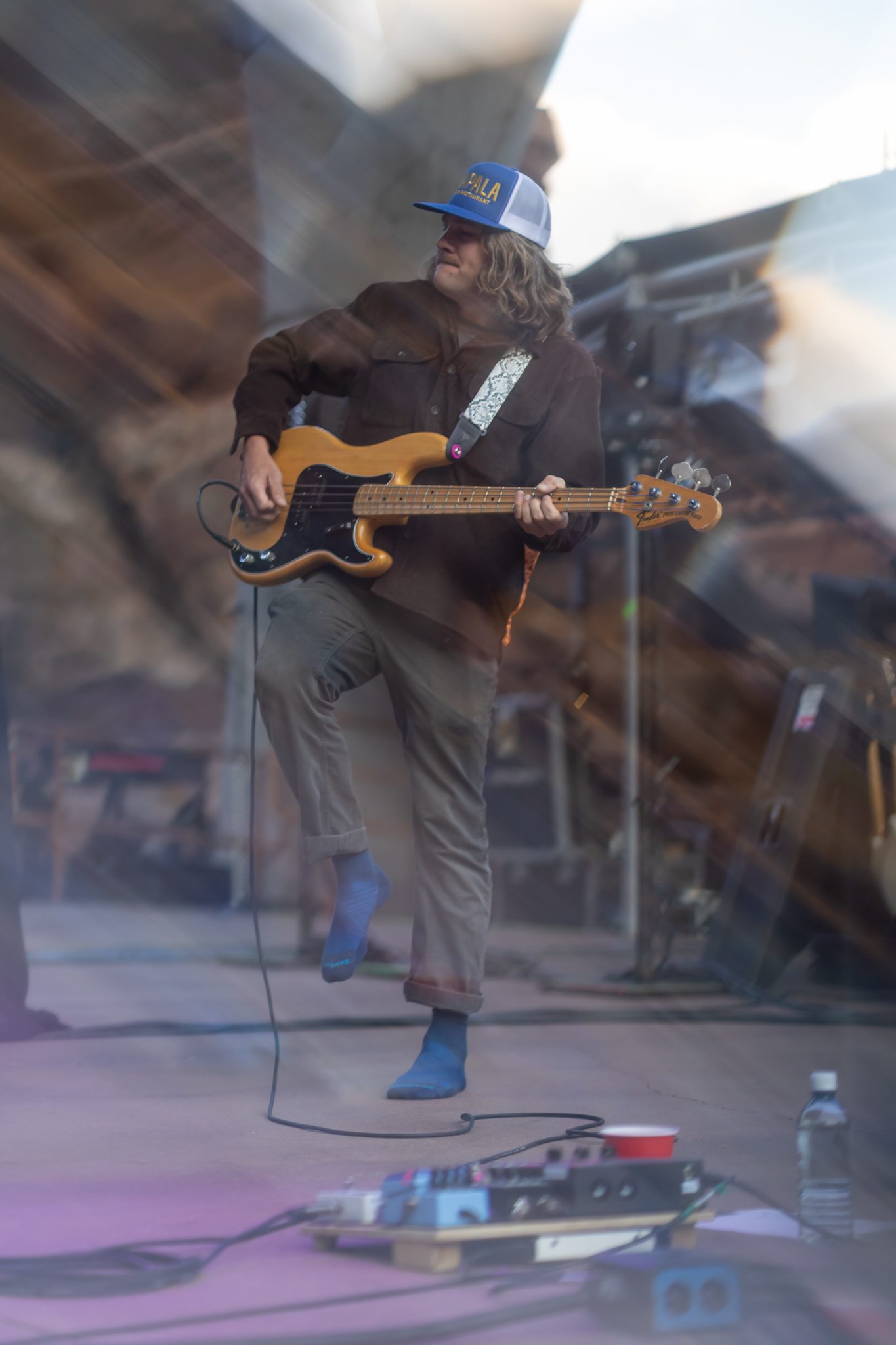 A musician playing a bass guitar on stage, wearing a baseball cap and standing on one leg, with a colorful background of stage equipment.