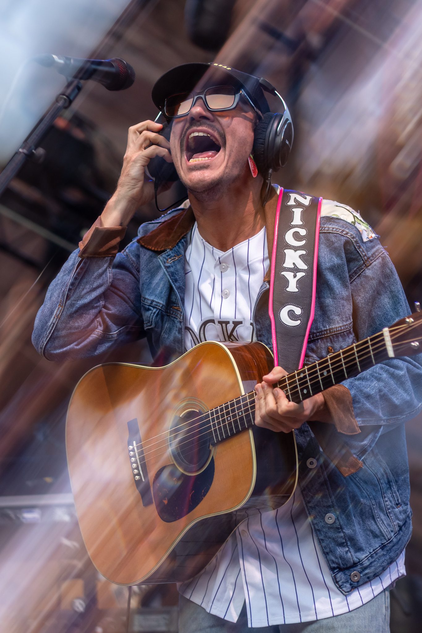 A musician passionately singing into a microphone while playing an acoustic guitar, wearing headphones and a denim jacket.