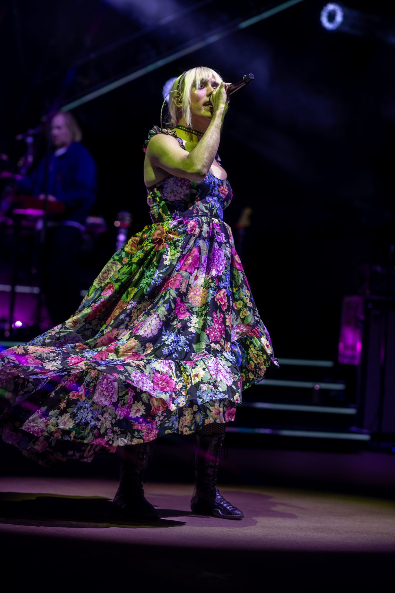 A singer performing on stage wearing a vibrant floral dress, passionately engaging with the audience under colorful stage lighting.