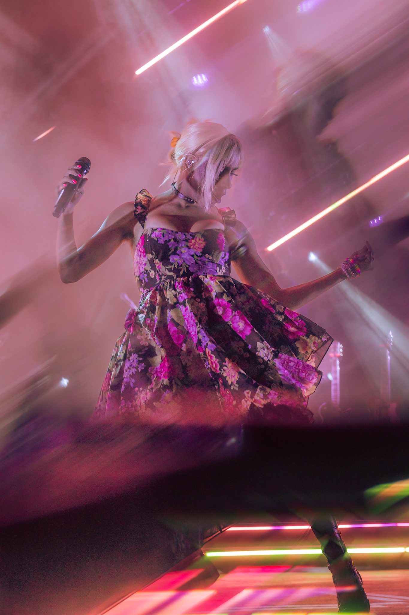 A performer on stage wearing a floral dress, holding a microphone, surrounded by vibrant lights and a smoky atmosphere, capturing the energy of a live concert.