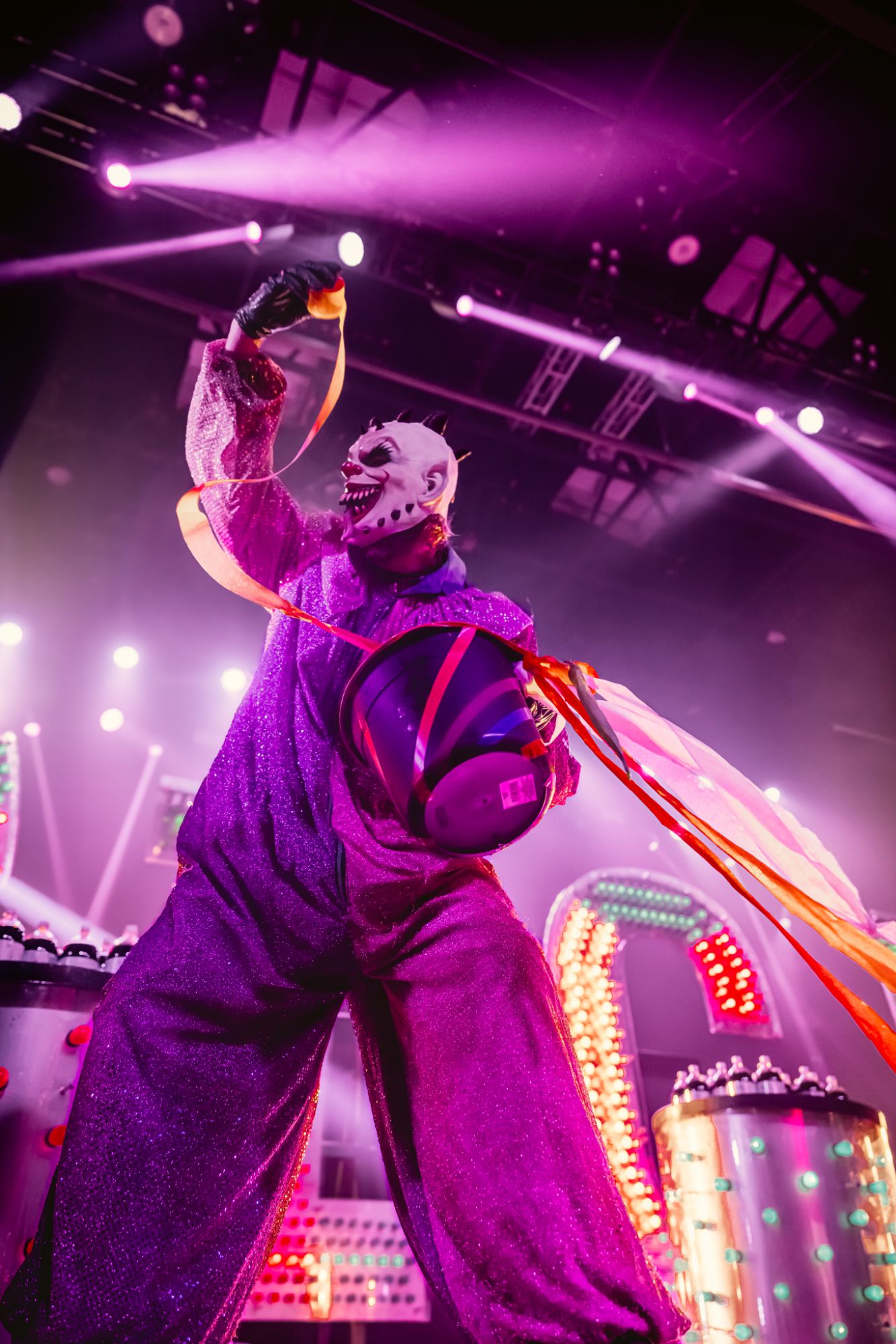 A performer in a purple glittery clown costume with a dramatic mask holds a colorful ribbon while on stage, surrounded by vibrant lights and theatrical props.
