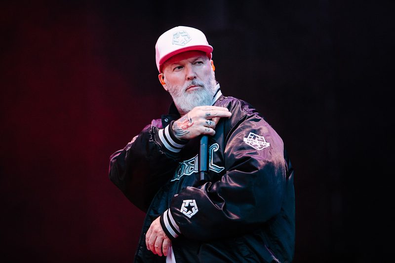 A musician on stage wearing a black jacket and a pink cap, looking intently into the crowd while holding a microphone, with colorful lighting in the background.