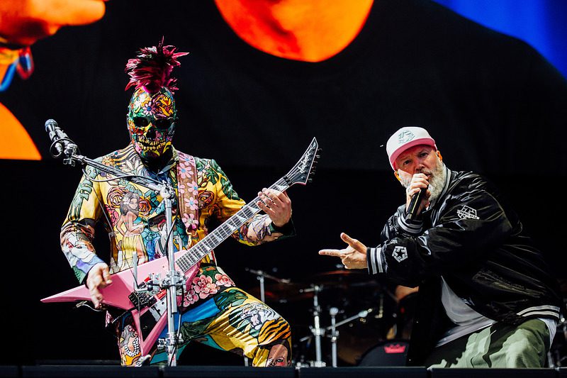 A vibrant live performance by a band, featuring a guitarist in a colorful outfit with intricate designs and a face and head covered in body paint, alongside a male vocalist in a black jacket and light-colored cap, engaging with the audience.