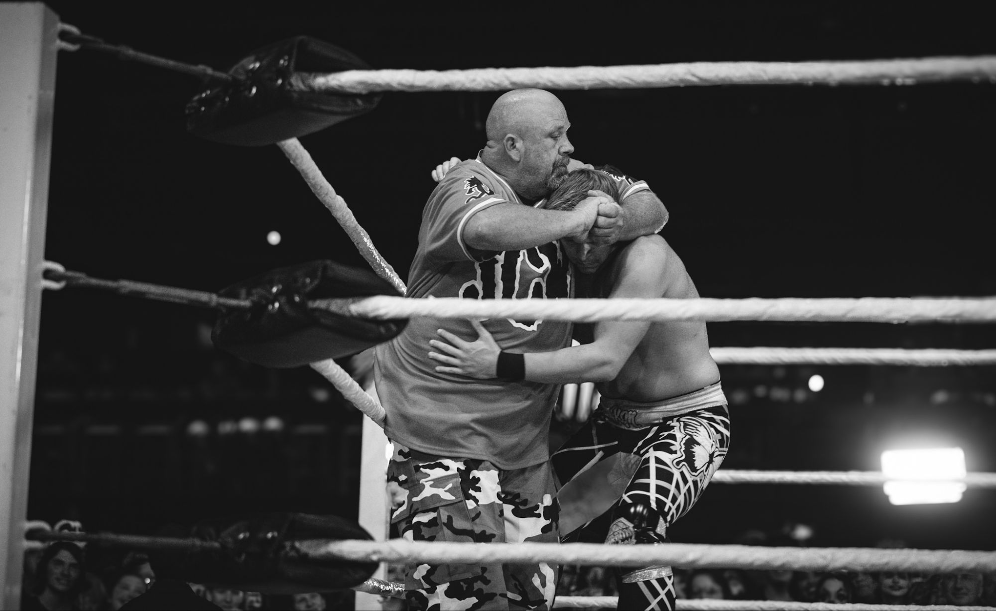 Two wrestlers are engaged in a hold inside the wrestling ring, with ropes and a cheering crowd visible in the background. One wrestler is wearing camouflage shorts and a colorful shirt, while the other has a muscular build and is dressed in patterned tights.