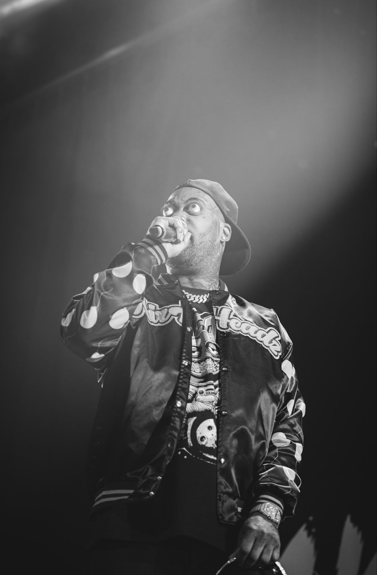 A rapper performing on stage, wearing a black jacket with polka dots and a cap, holding a microphone, with expressive facial features and dramatic lighting.