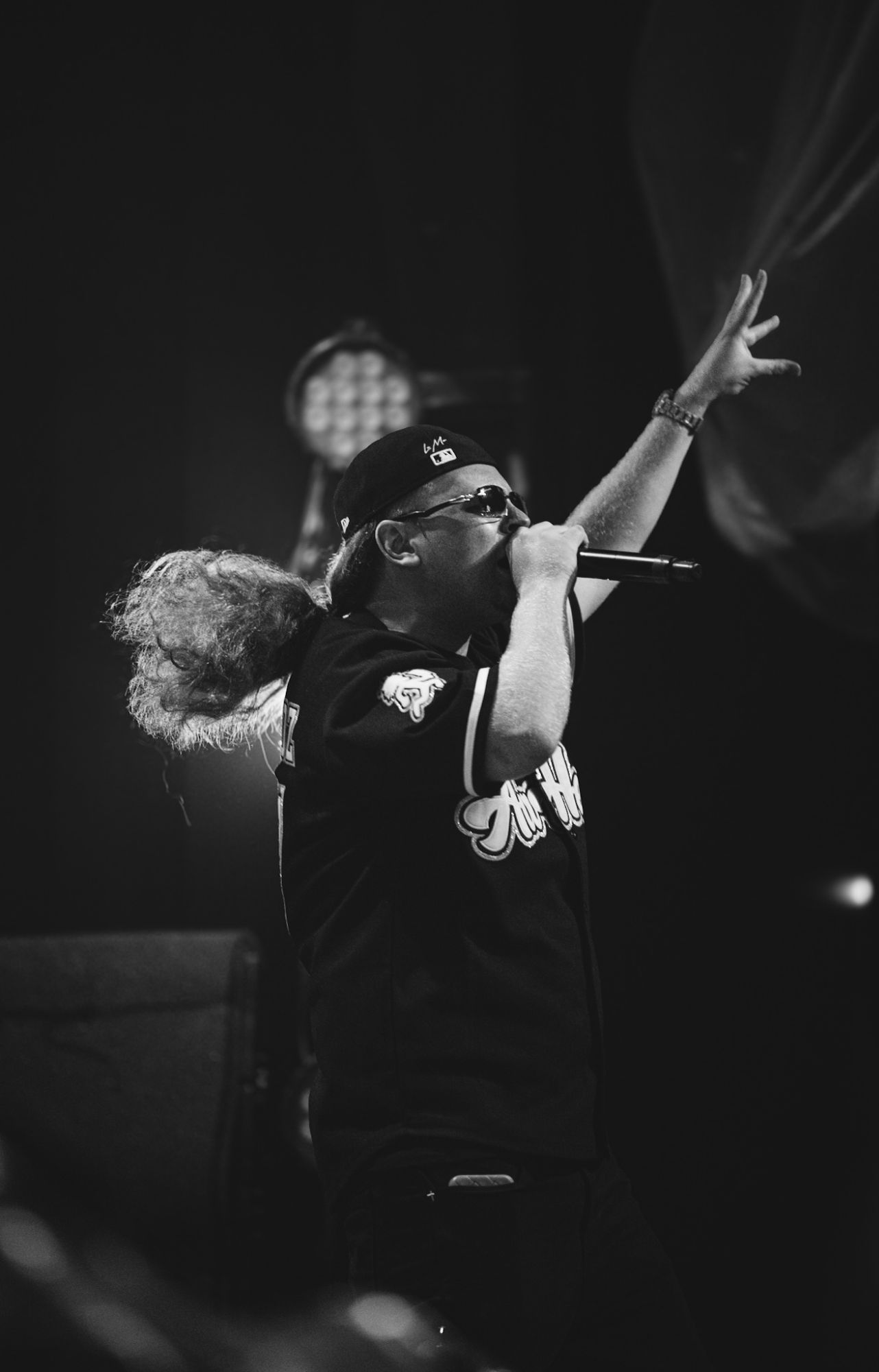 A performer passionately singing into a microphone while raising one hand, wearing sunglasses and a baseball cap, amidst dramatic stage lighting.