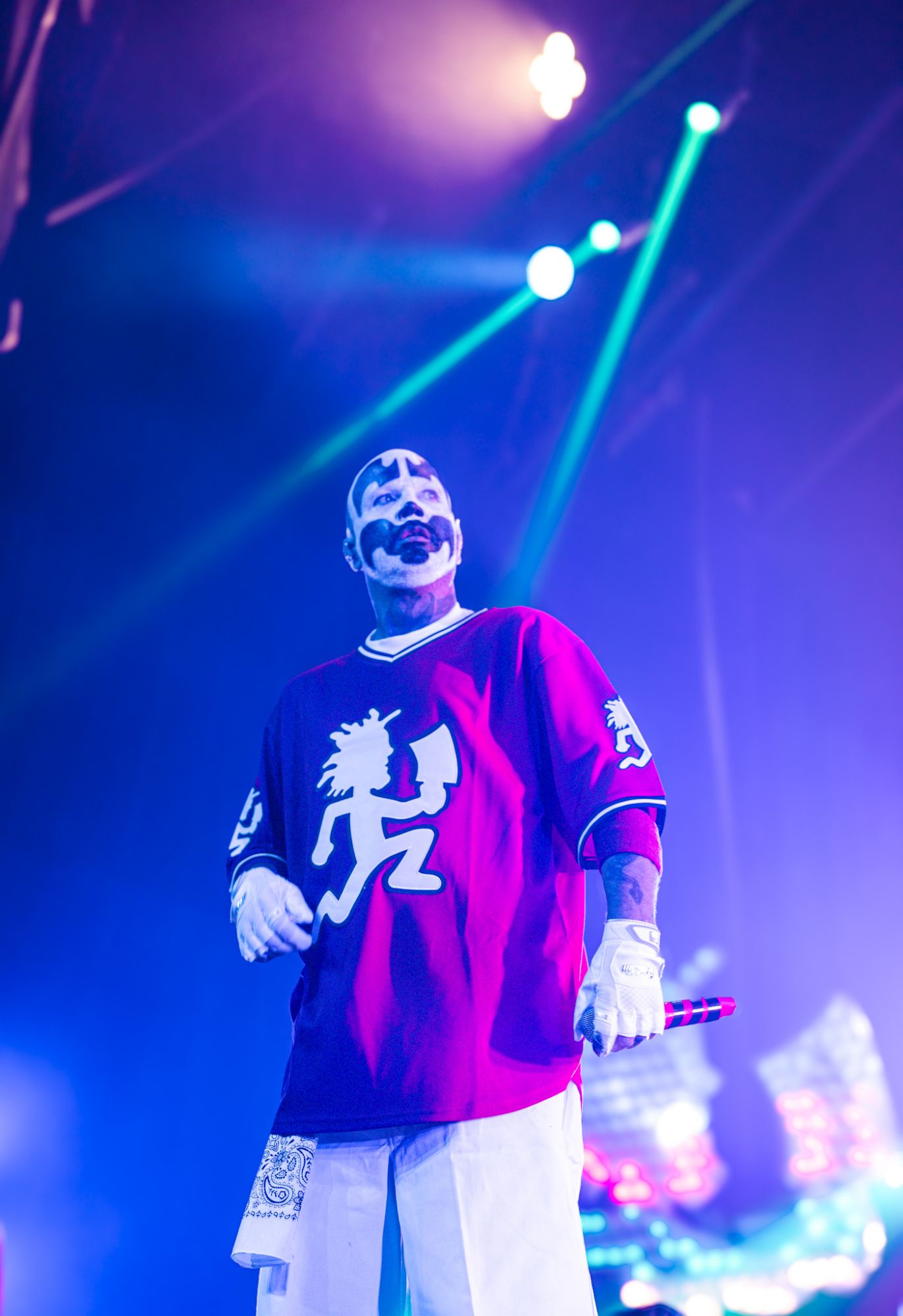 A performer wearing a red jersey featuring a Juggalo logo stands on stage, holding a microphone, with colorful stage lights in the background.