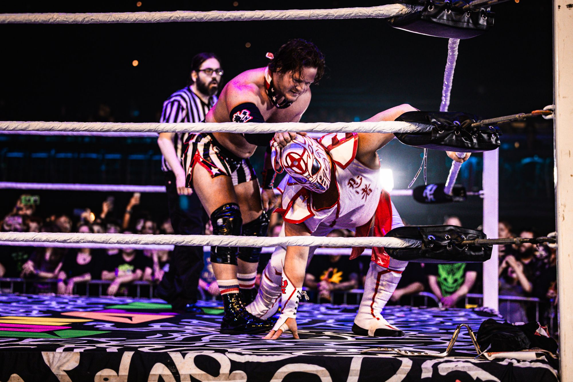 Two wrestlers in a ring, one applying pressure to the other while a referee observes. The crowd is visible in the background.