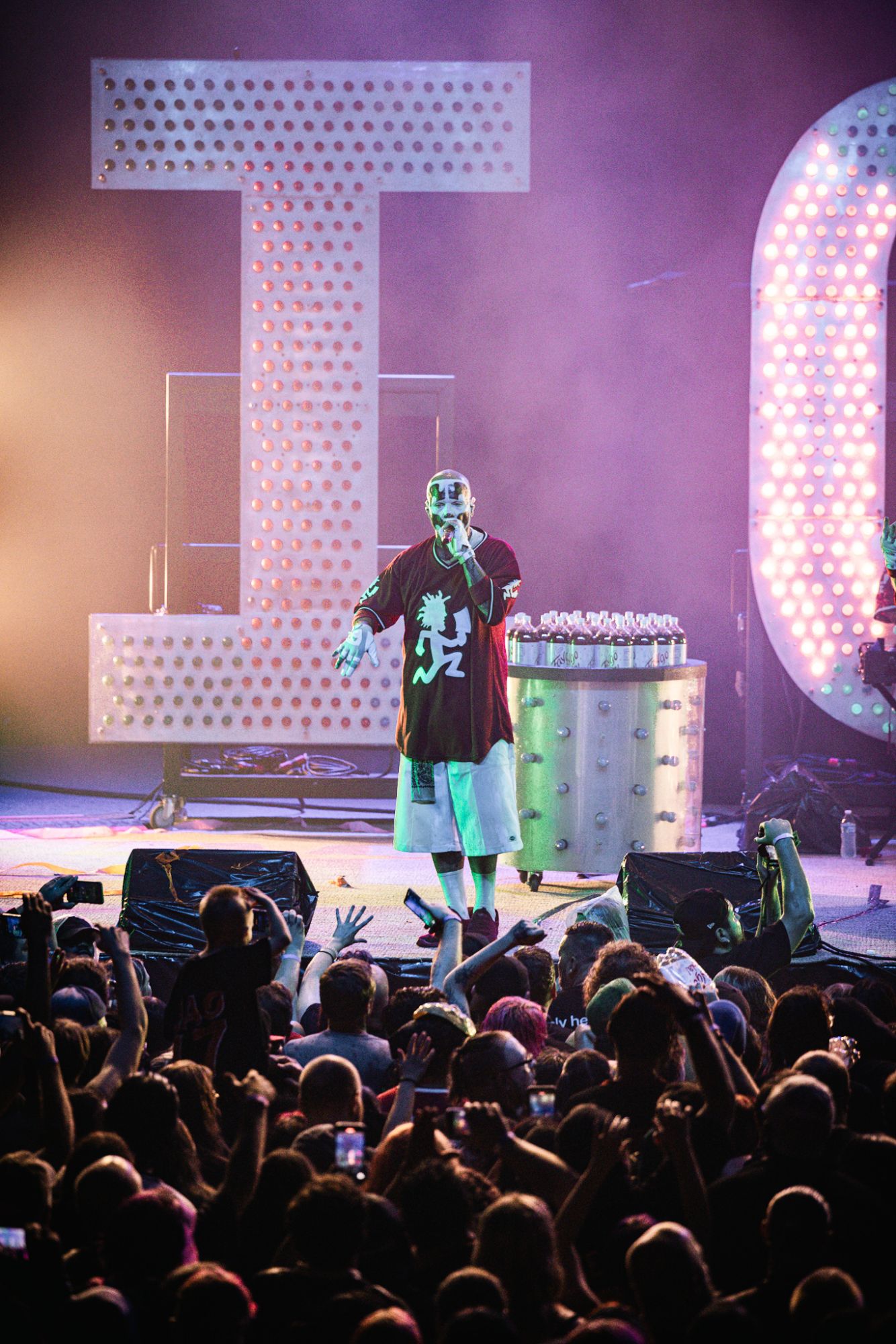 A performer wearing a red shirt and shorts is on stage, engaging with the crowd during a concert. Large illuminated letters are visible in the background, and fans are reaching out toward the artist.
