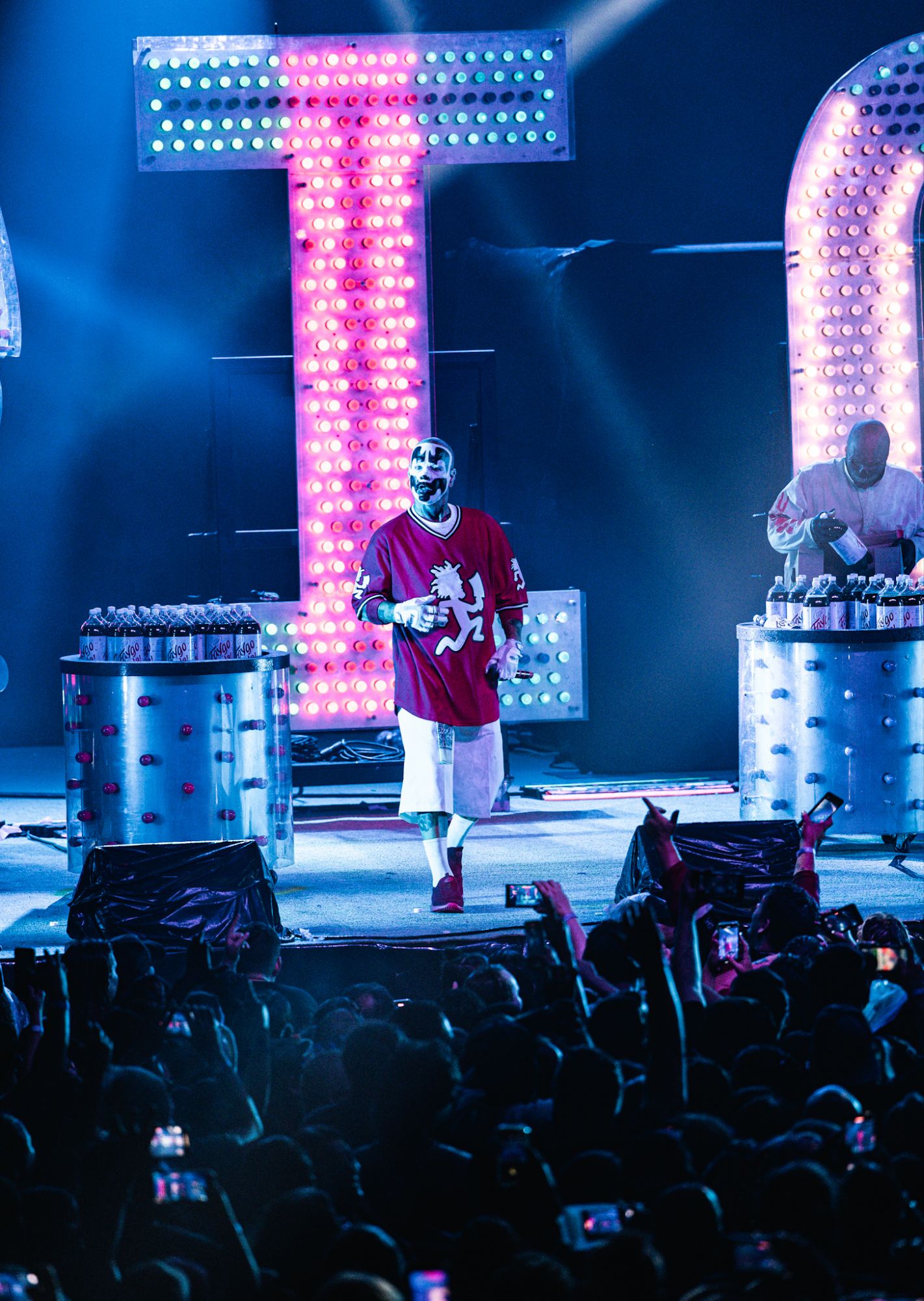 A performer from Insane Clown Posse on stage, dressed in a red jersey and white shorts, interacting with the audience. Colorful lights and soda cans are visible in the background, creating a vibrant concert atmosphere.