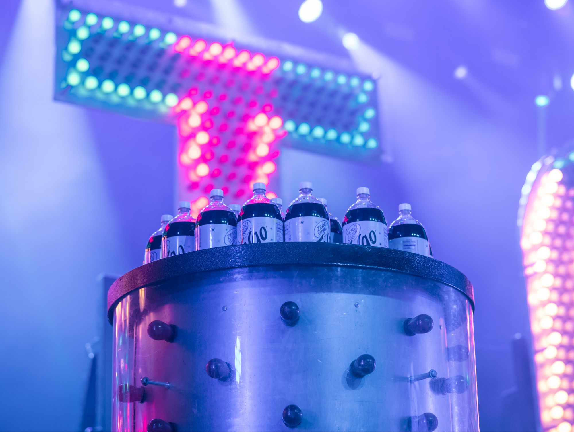 Close-up of bottles of soda on a stage, illuminated by colorful lights in a concert setting.