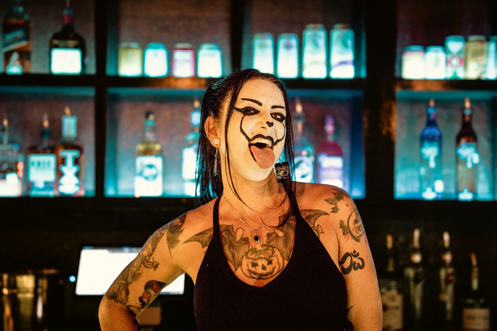 A woman with face paint resembling a clown sticking out her tongue, standing in front of a brightly lit bar with various bottles in the background.