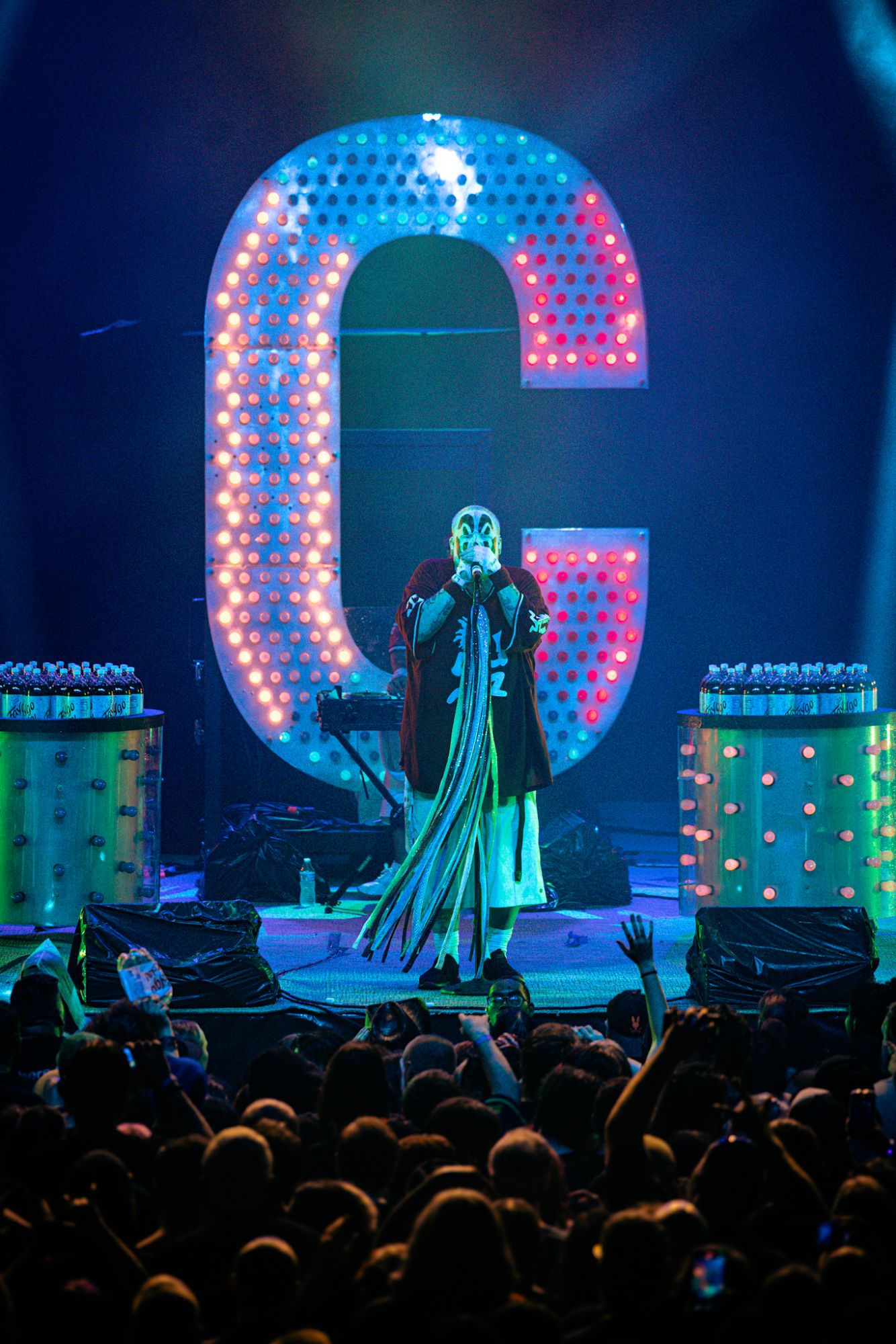 A performer in a clown costume with colorful face paint, holding a microphone and standing in front of a large illuminated letter 'C' on stage during a concert, with a lively crowd raising their hands in the foreground.