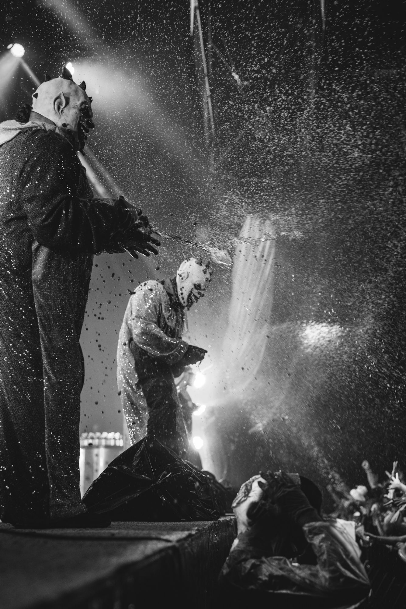 Two performers in clown costumes on stage, spraying the audience with colorful liquids during a high-energy performance, with bright stage lights and an enthusiastic crowd visible.