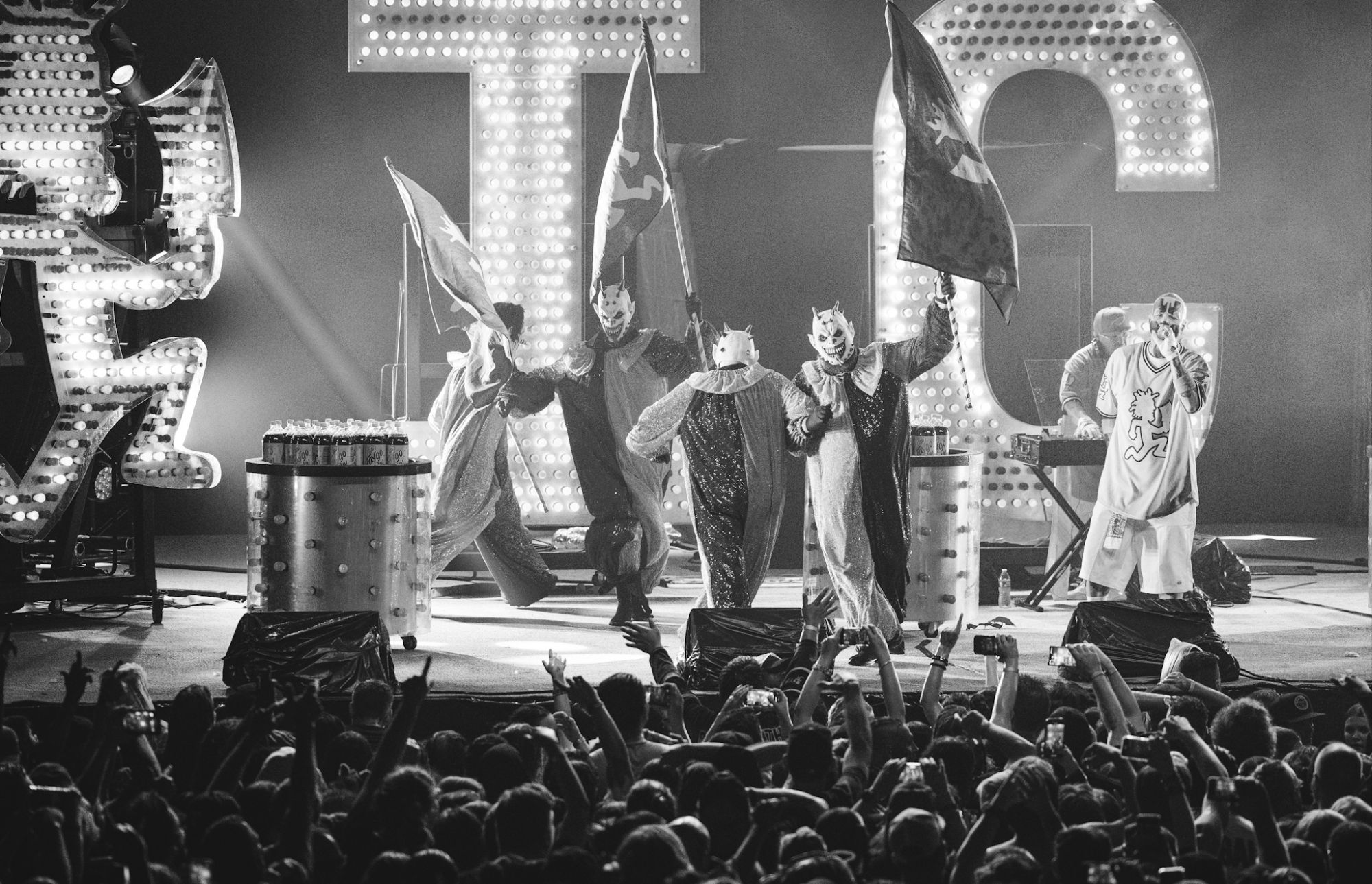 Insane Clown Posse performing on stage, dressed in colorful clown outfits and masks, holding flags as the crowd cheers enthusiastically.