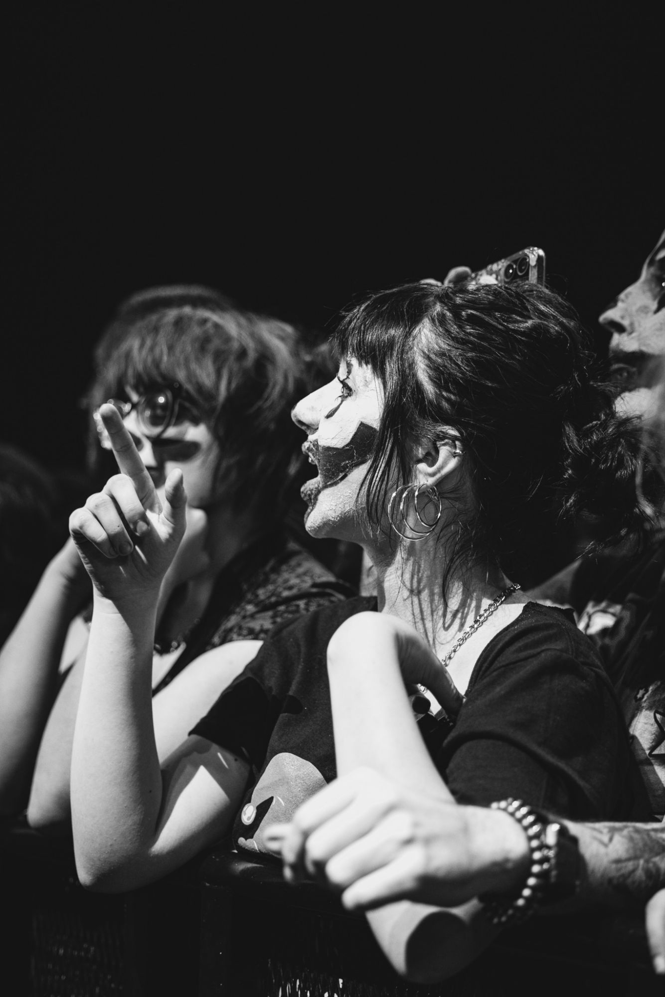 A close-up of excited fans at a concert, showing a woman with face paint and expressive gestures, while others with distinctive hairstyles and accessories are behind her.
