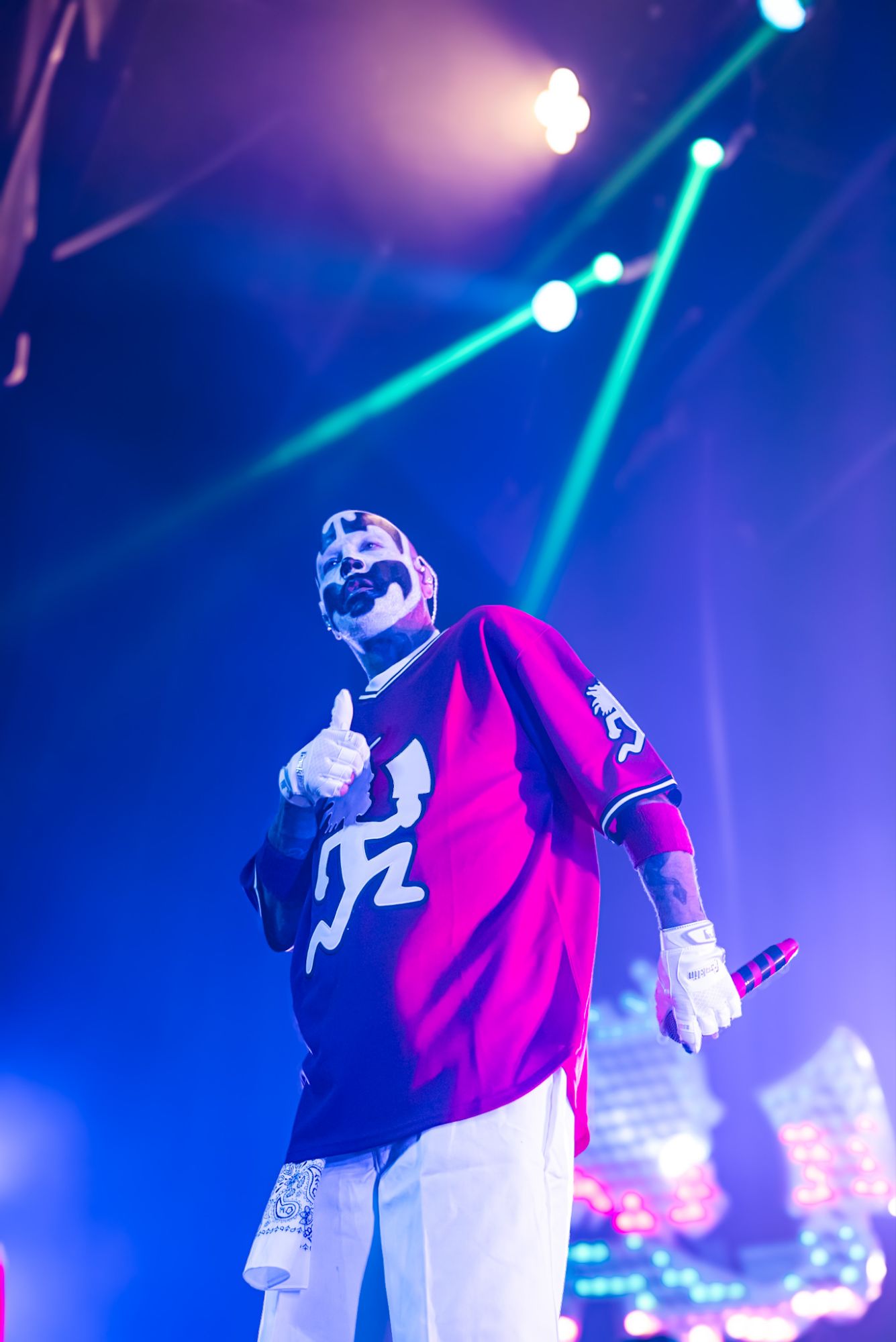 A performer from Insane Clown Posse on stage, wearing a red jersey with white gloves, giving a thumbs up against a backdrop of colorful stage lights.