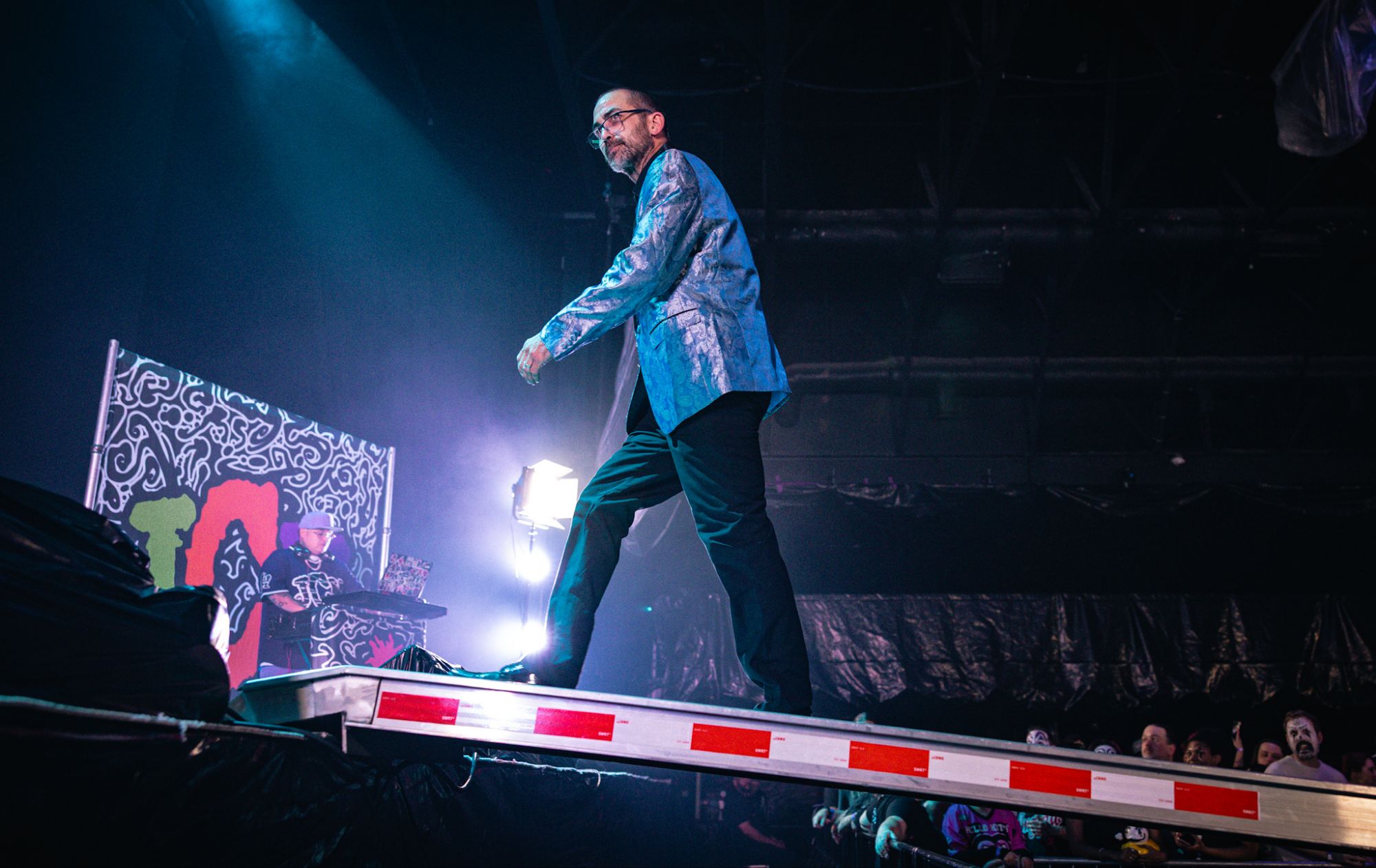 A performer in a shiny blue jacket walks across a narrow platform on stage, with vibrant lighting and a DJ in the background. The audience watches eagerly from below.