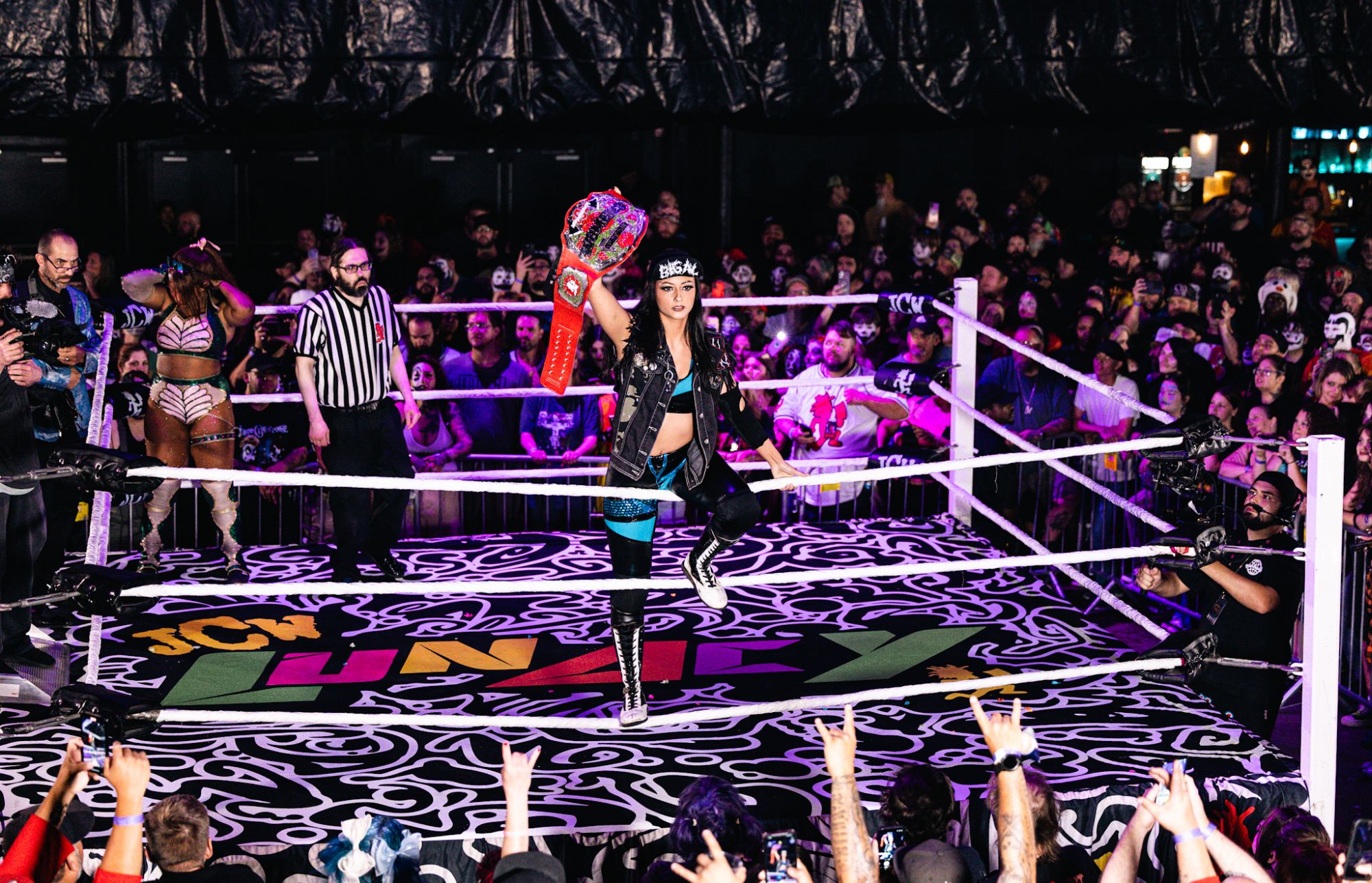 A female wrestler celebrates with a championship belt while the crowd cheers in a wrestling ring at a lively event.
