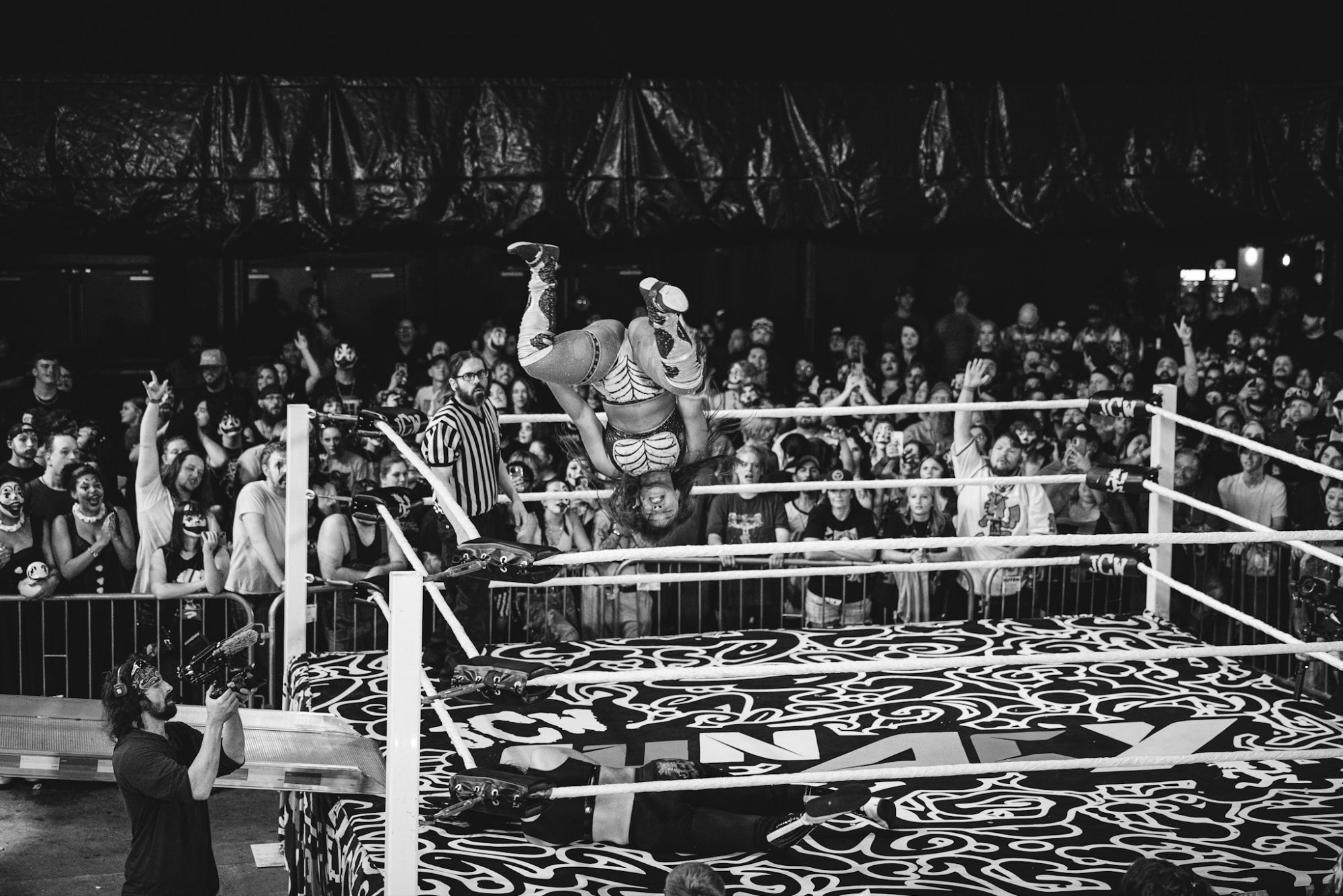 A wrestler performs a high-flying move above the ring during a match, surrounded by an enthusiastic audience, some wearing Juggalo face paint.