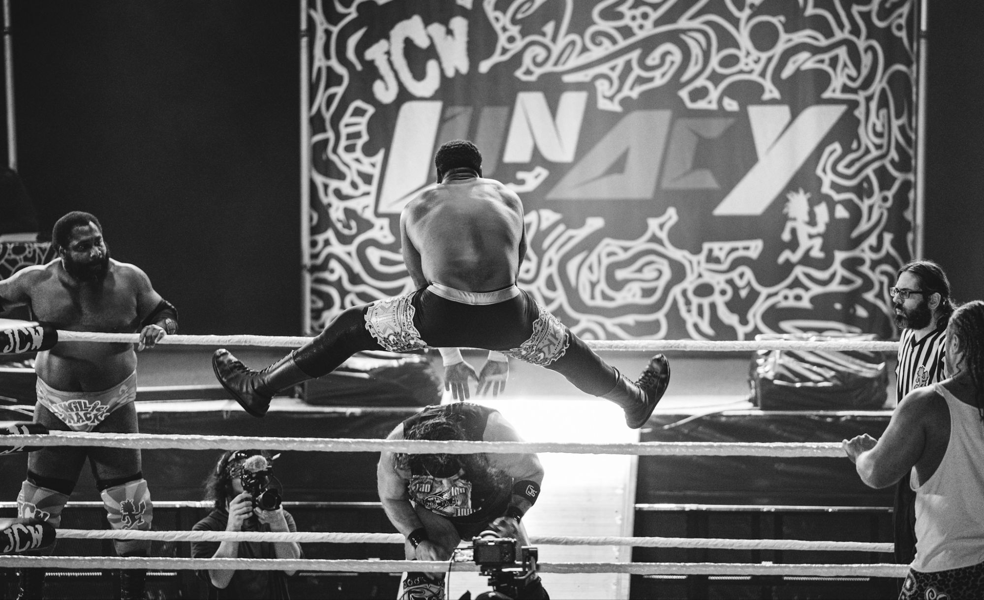 A wrestler leaps off the ropes in an action-packed moment during a JCW wrestling match, with spectators and wrestlers focused on the ring.