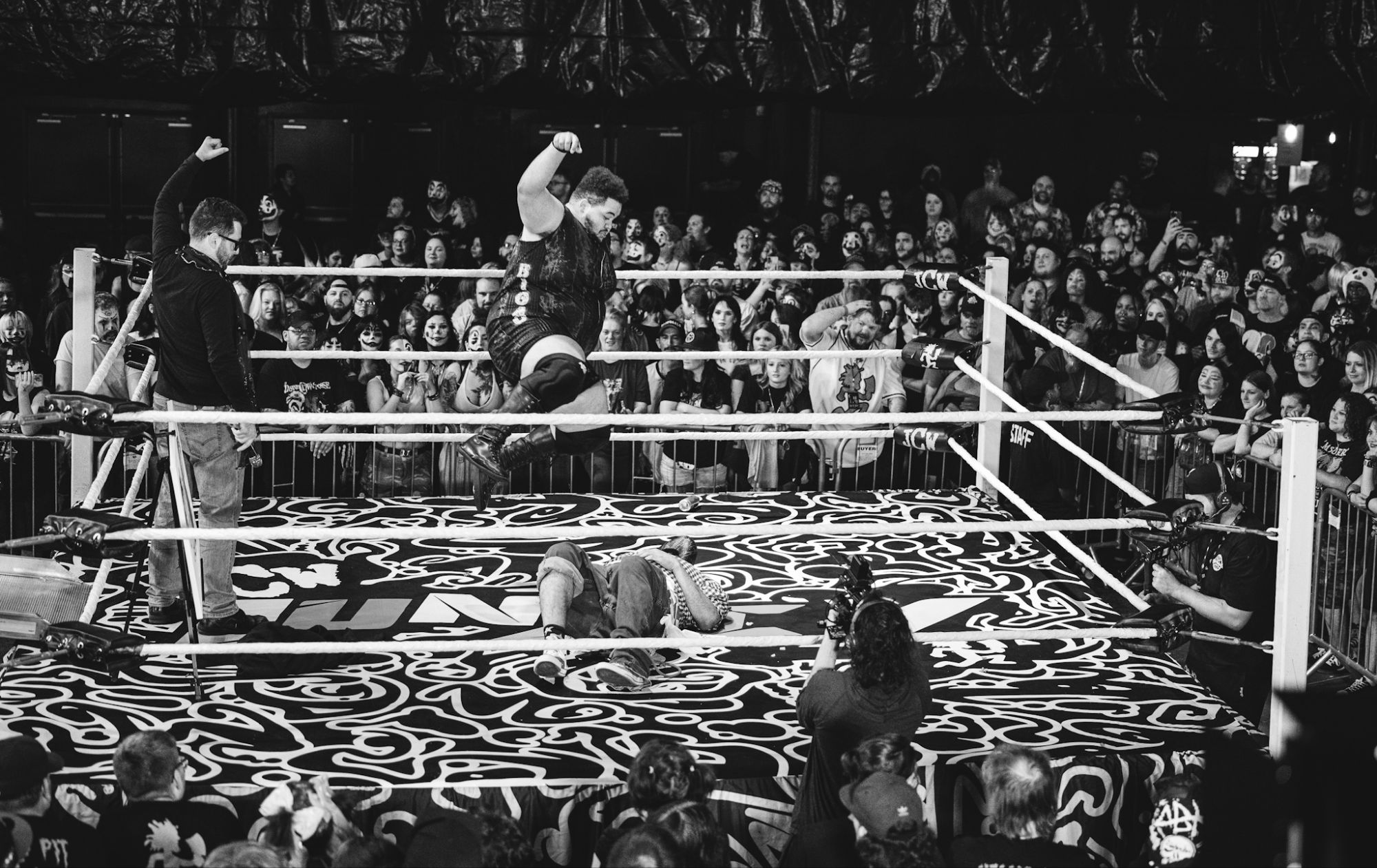 A wrestler leaping off the ropes above an opponent lying on the mat during a live wrestling event, with a cheering crowd in the background.