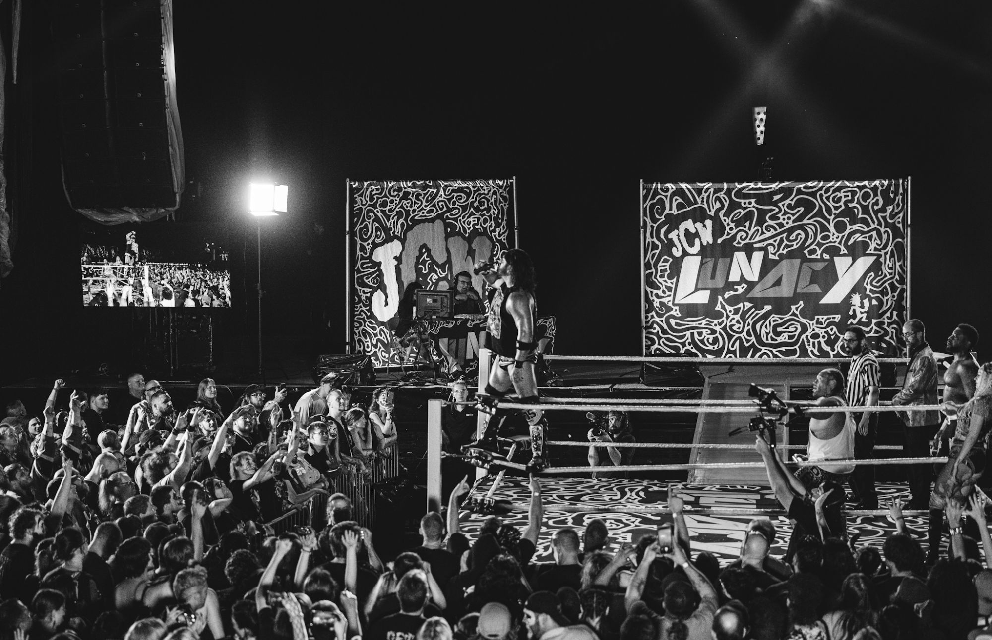 Black and white image of a wrestling ring with a performer standing on the ropes, surrounded by an enthusiastic audience. Behind the ring are colorful banners displaying 'JCW LUNACY.'
