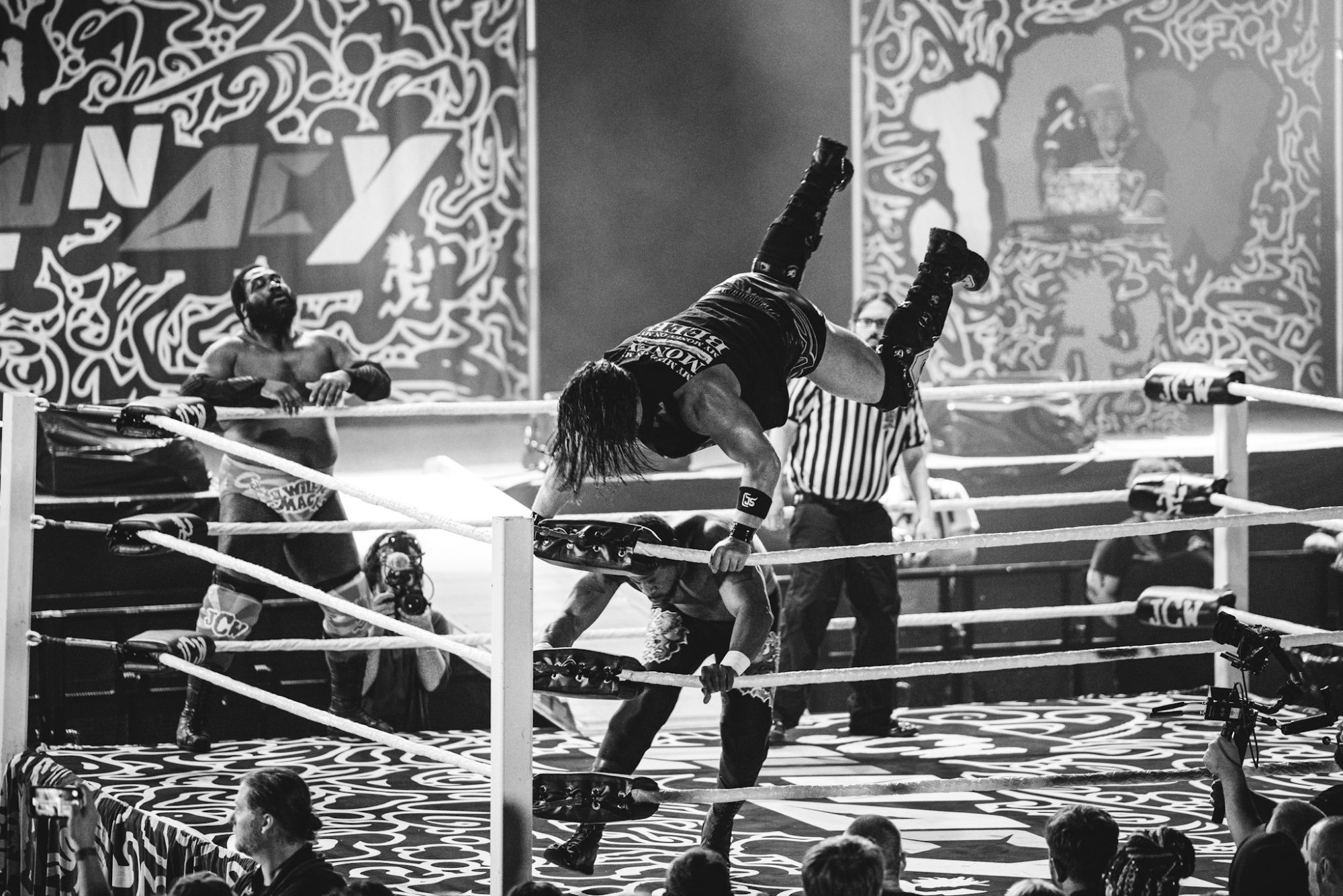 A wrestling match featuring two competitors in action within a ring, surrounded by an energetic audience and vibrant, patterned backdrop.