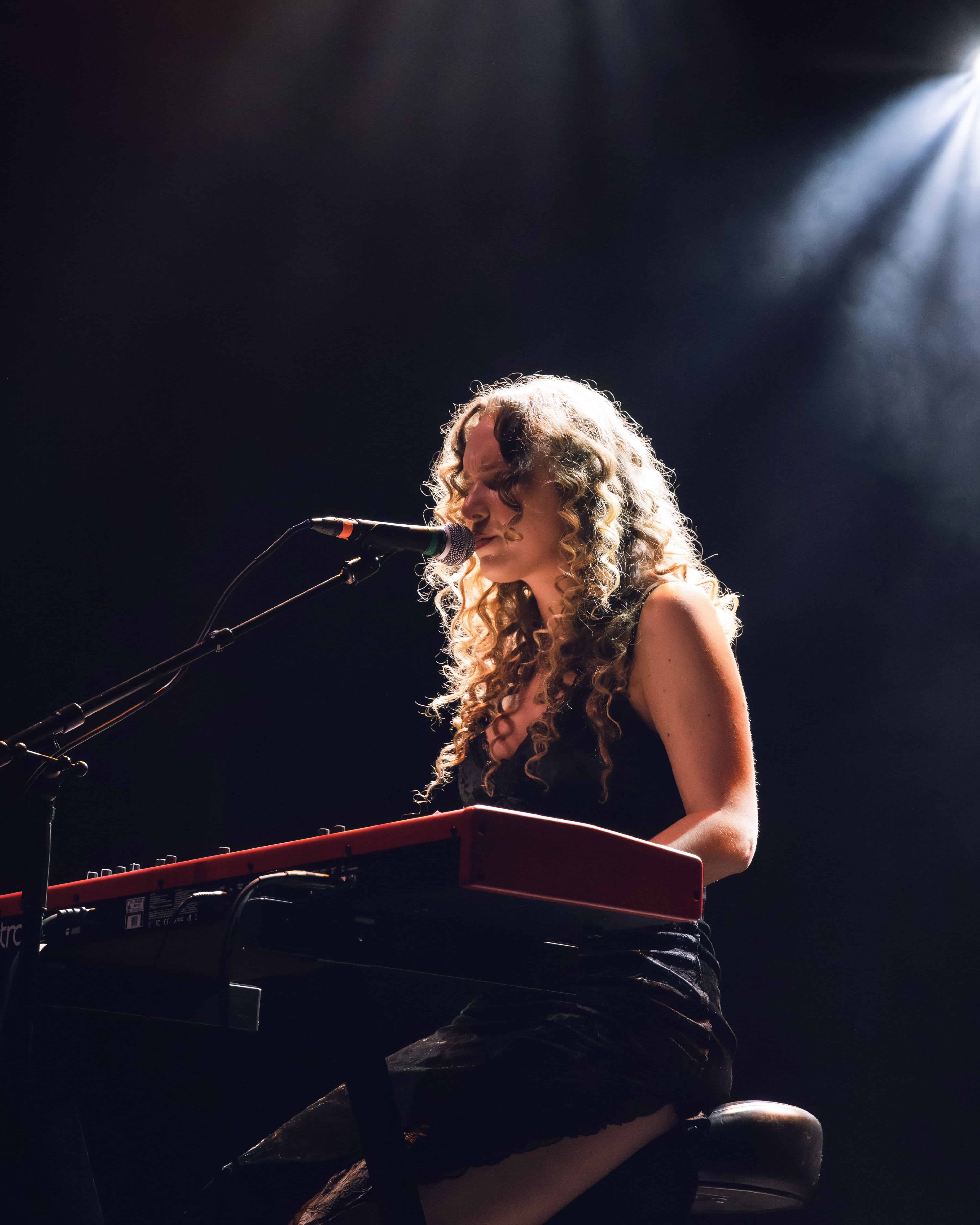 A singer with curly hair performing on stage, playing a red keyboard, with dramatic lighting highlighting her presence.