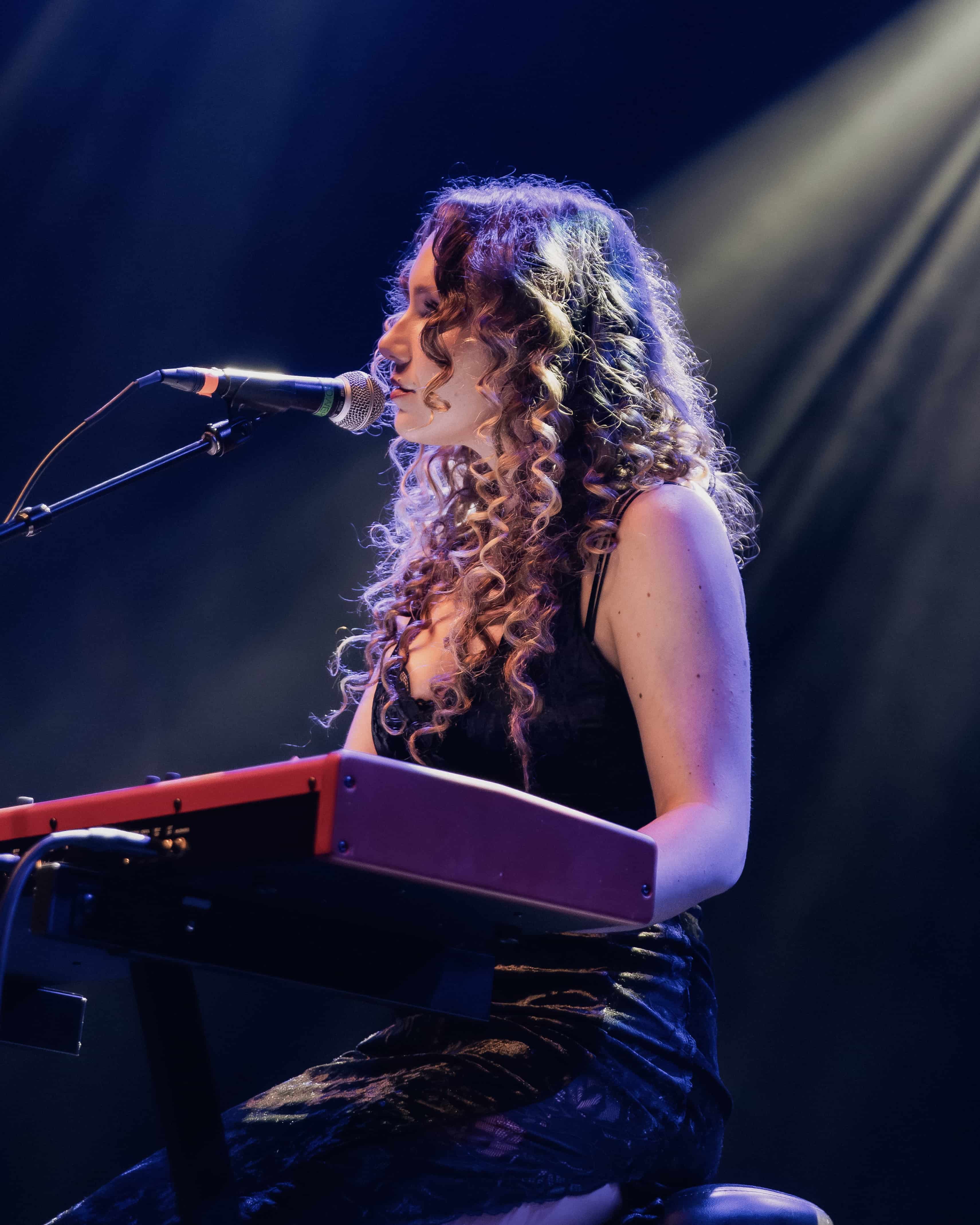 A female musician with curly hair performs at a microphone while playing a keyboard, illuminated by stage lights.