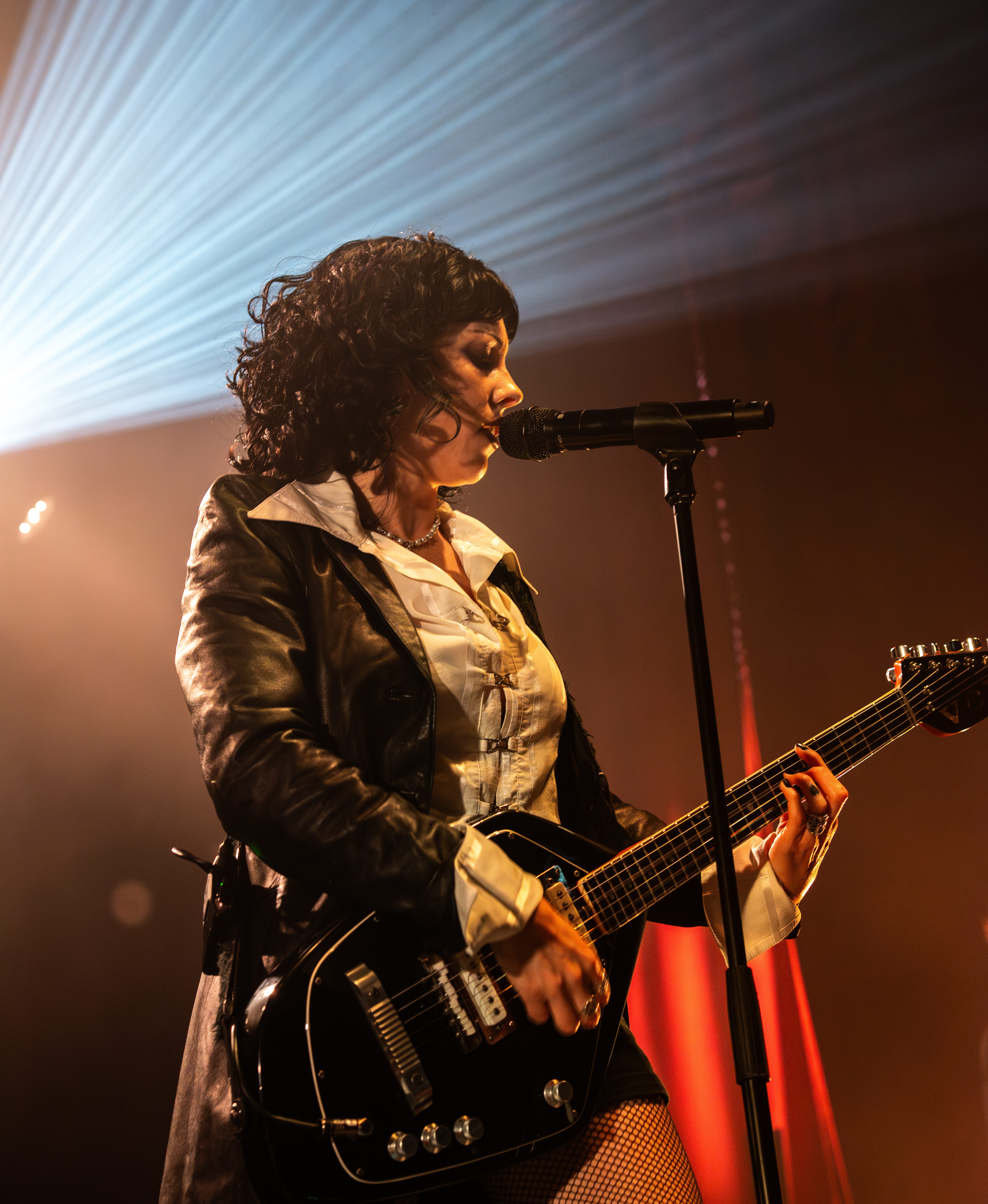 A female musician performs on stage, holding a black guitar and singing into a microphone, with dramatic stage lighting in the background.