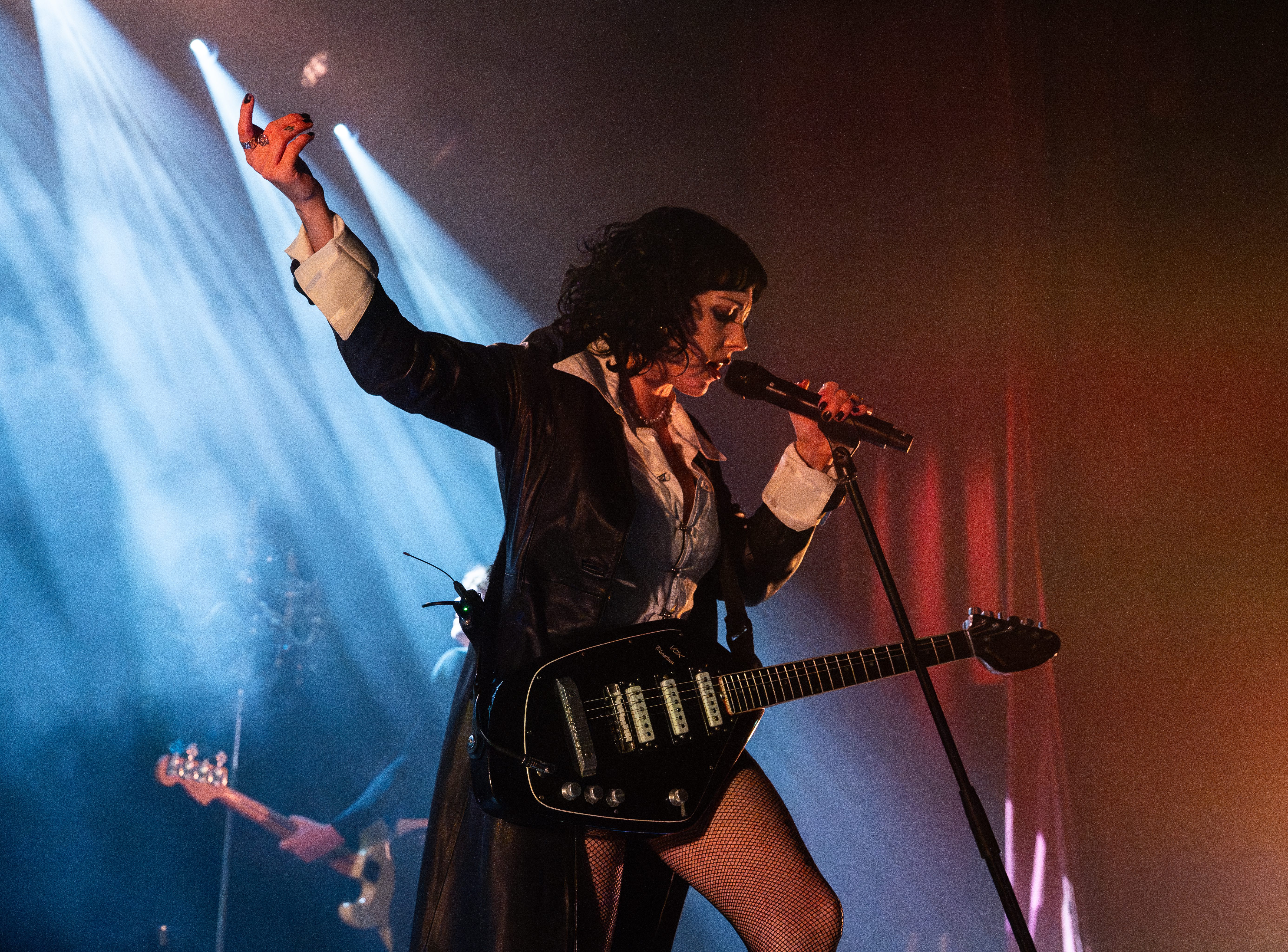 A performer on stage passionately singing into a microphone while playing a guitar, wearing a stylish outfit with dramatic lighting in the background.