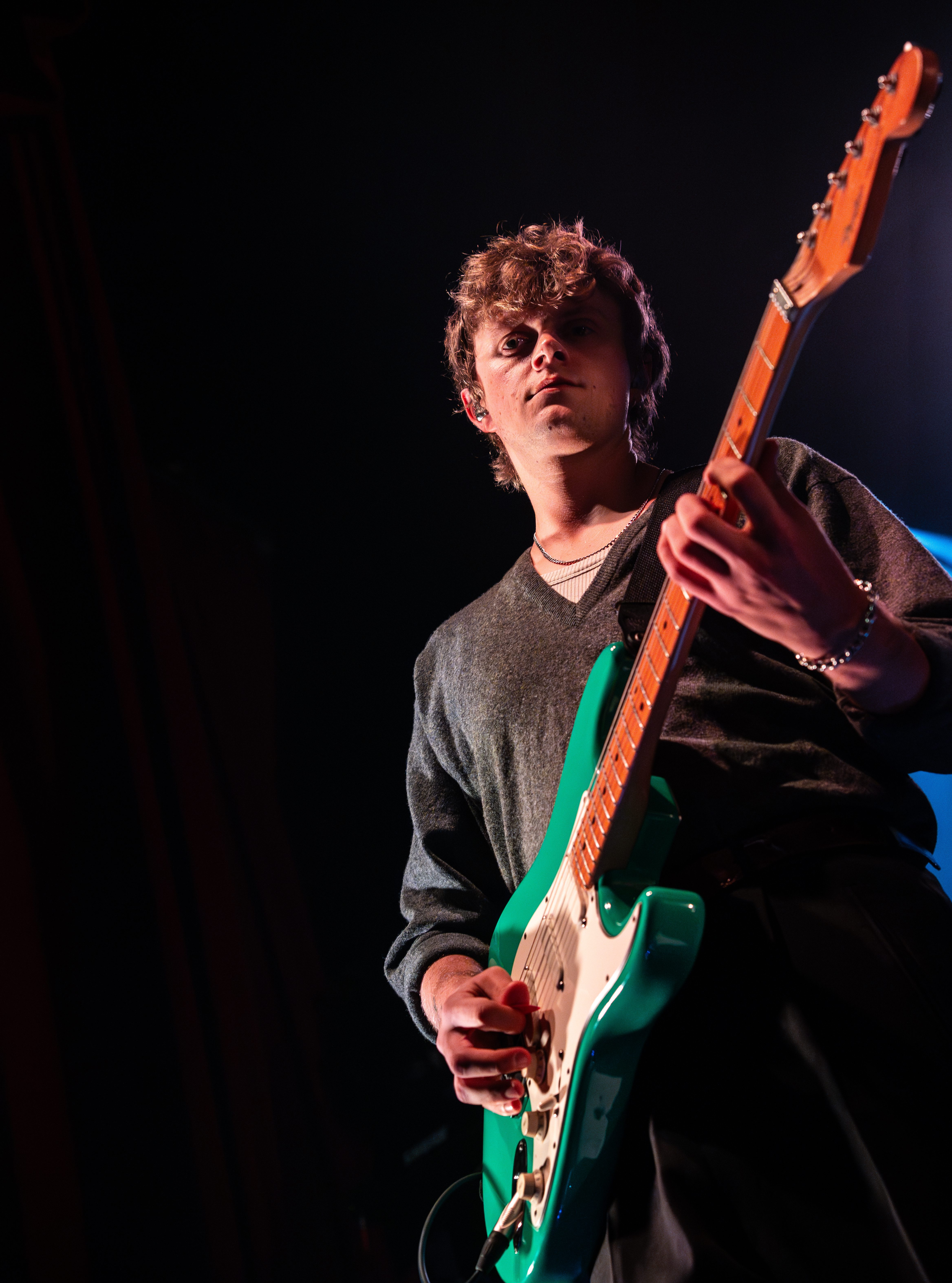 A musician performing on stage, holding a green electric guitar with a focused expression. The setting is dimly lit, highlighting the performer's presence.