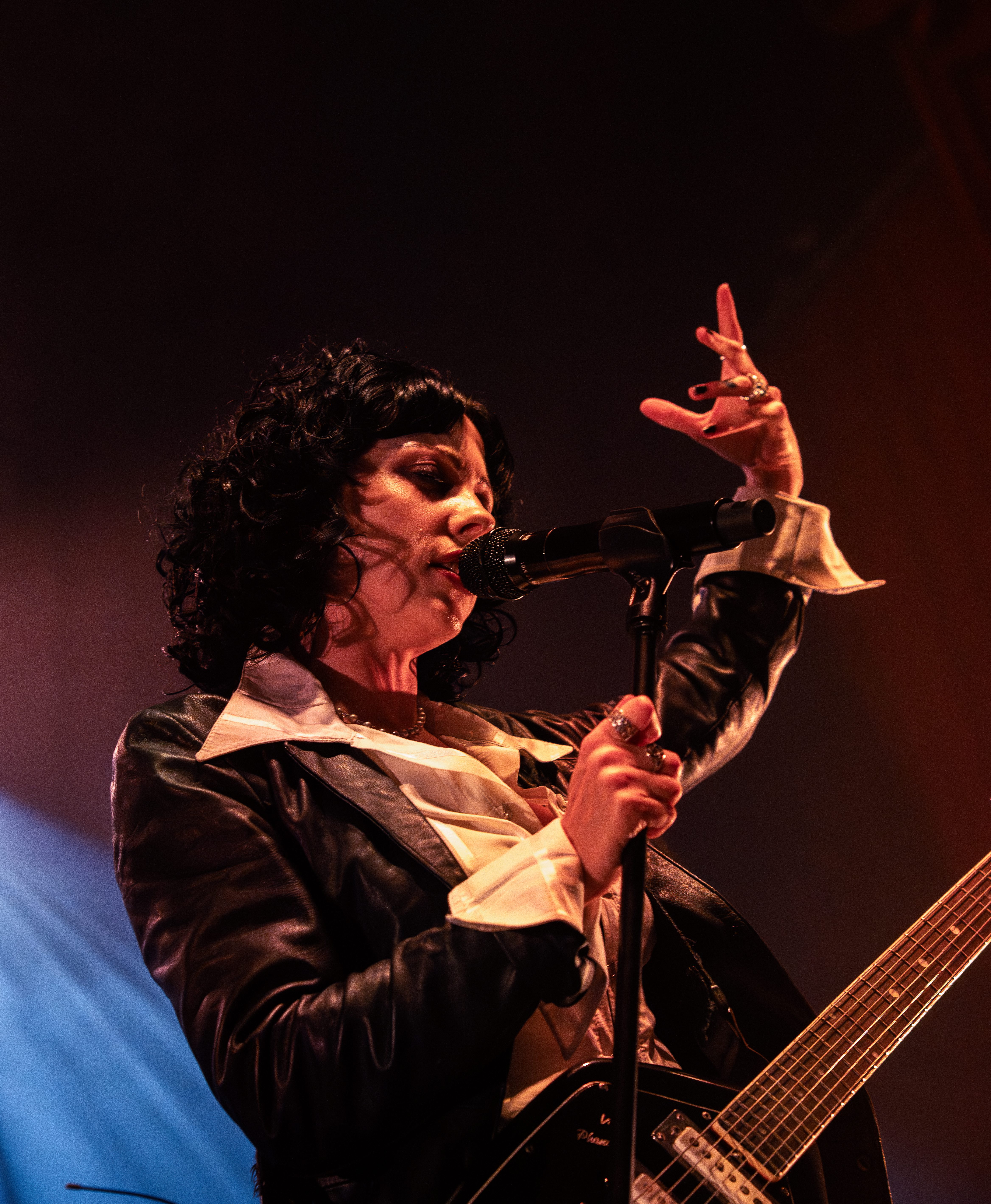 A female musician with curly hair performs passionately with a microphone in hand, wearing a stylish outfit that includes a leather jacket and a white shirt. She is animatedly gesturing with her free hand during a concert.
