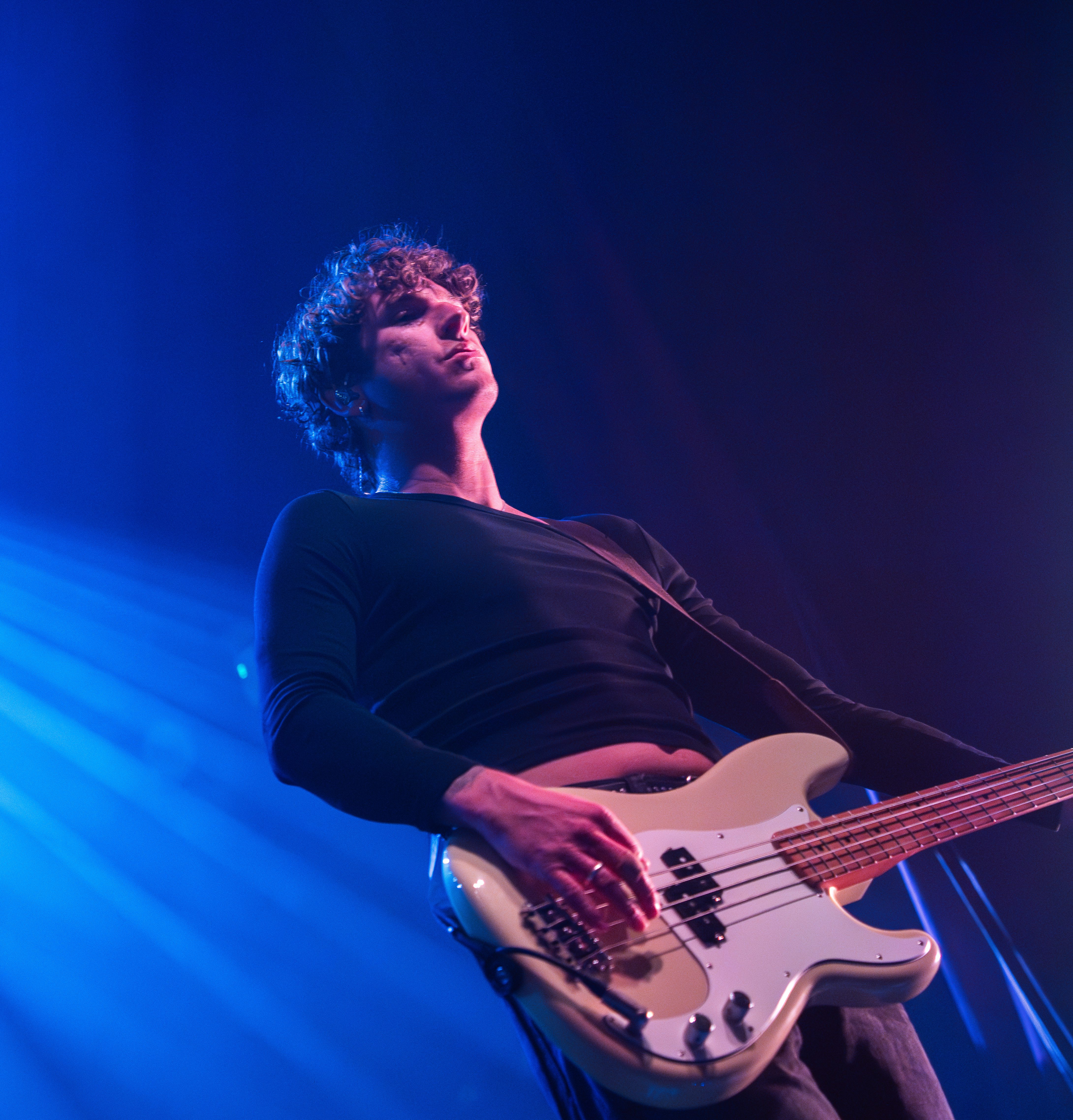 A male musician playing a bass guitar on stage, illuminated by blue stage lights.
