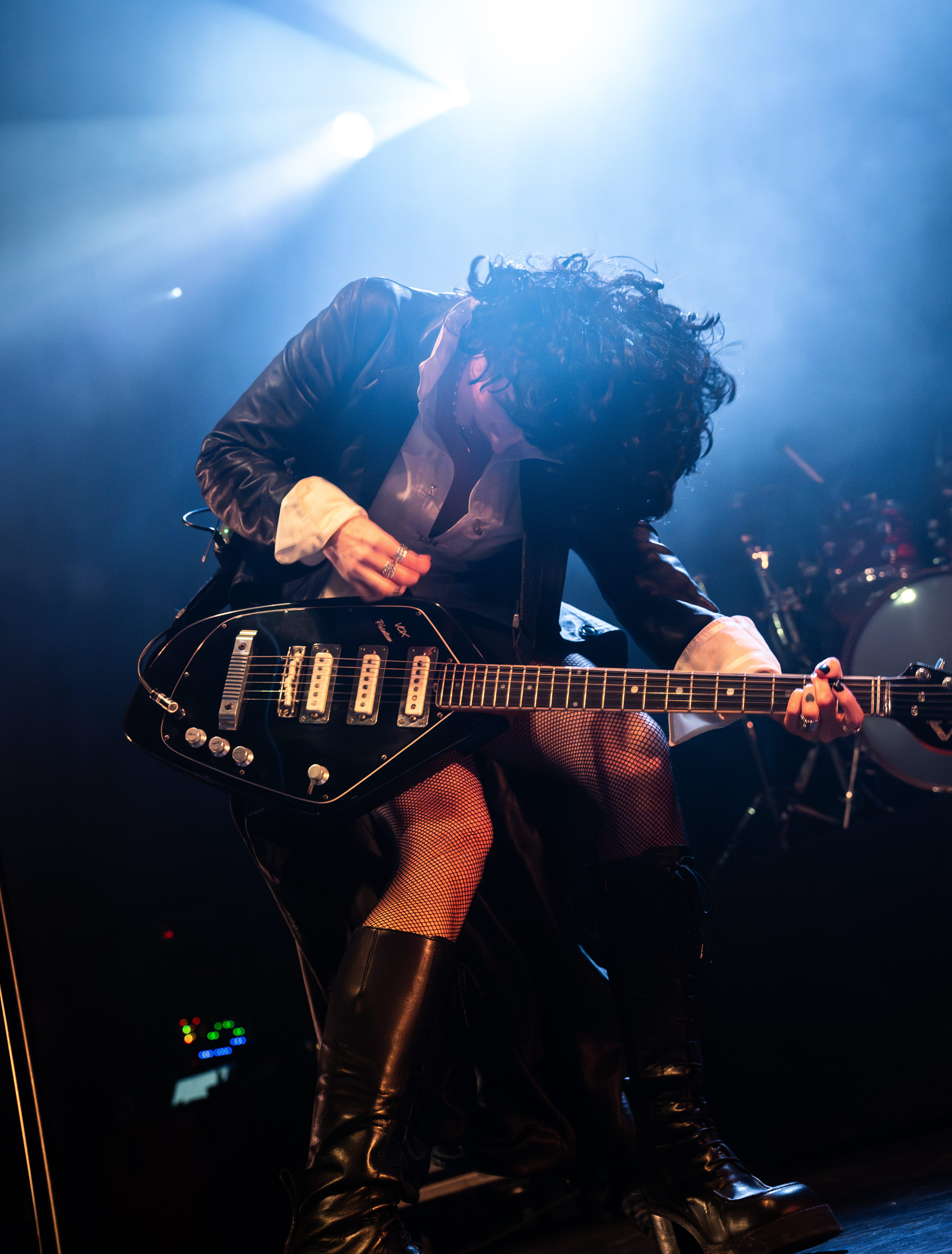 A musician performing on stage, wearing a black jacket and fishnet stockings, playing a black electric guitar under dramatic lighting.