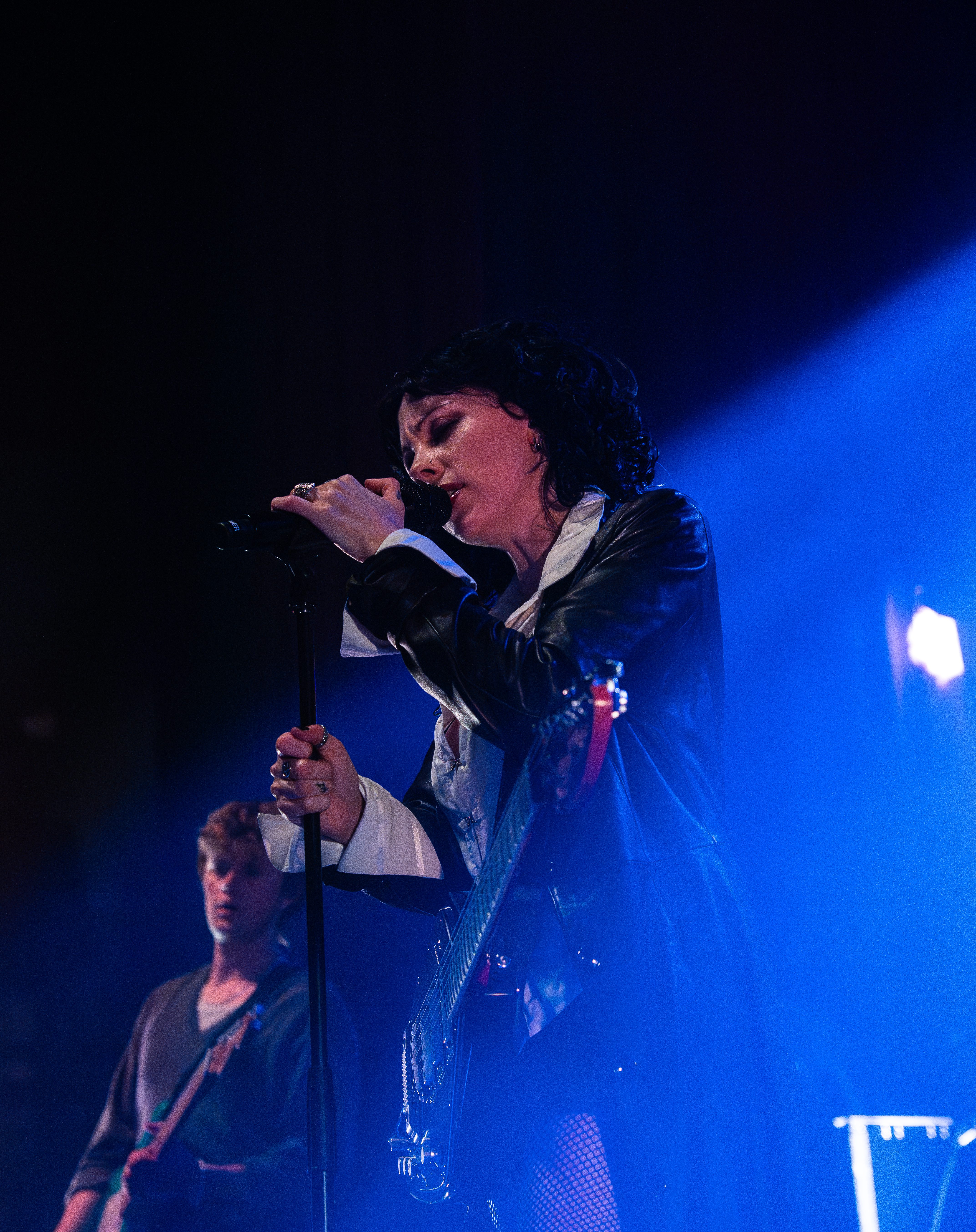 A female vocalist passionately performing on stage with a microphone in hand, wearing a leather jacket. A male musician playing bass is visible in the background under blue stage lighting.