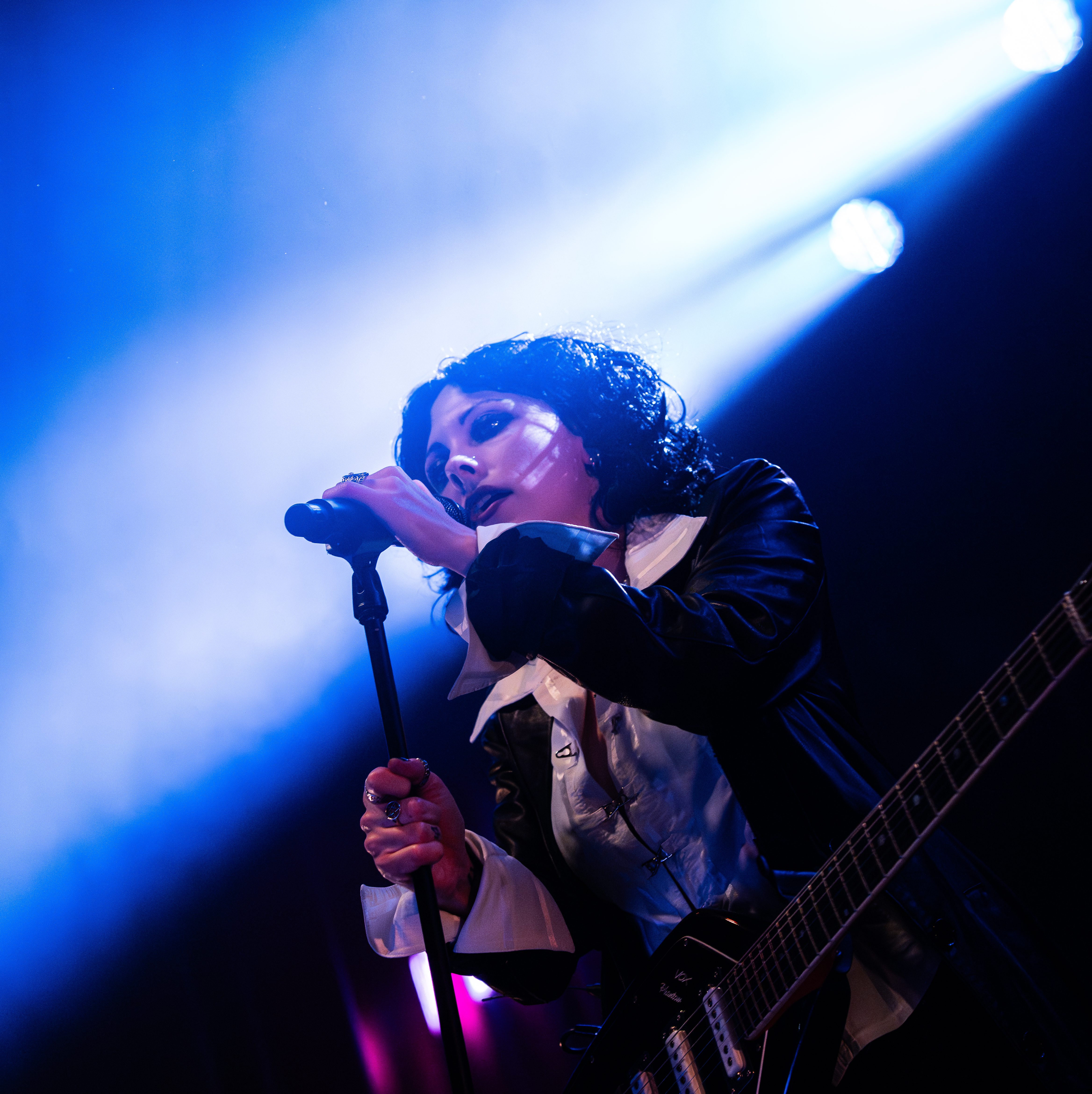 A singer performing on stage with a microphone, wearing a black jacket and a white shirt, illuminated by dramatic blue and pink lighting.