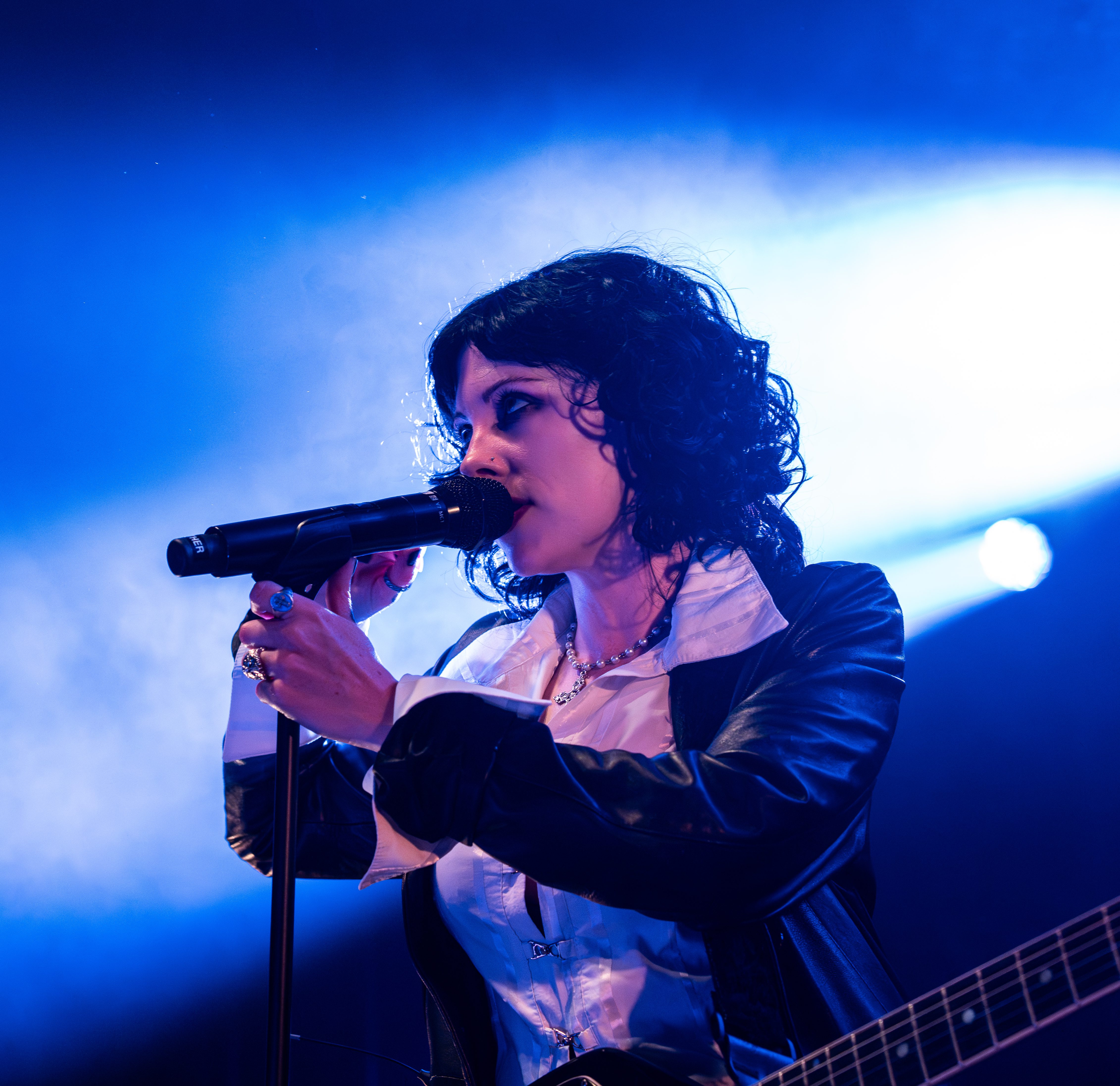 A singer holding a microphone on stage, dressed in a black leather jacket and a white shirt, with dramatic blue lighting in the background.