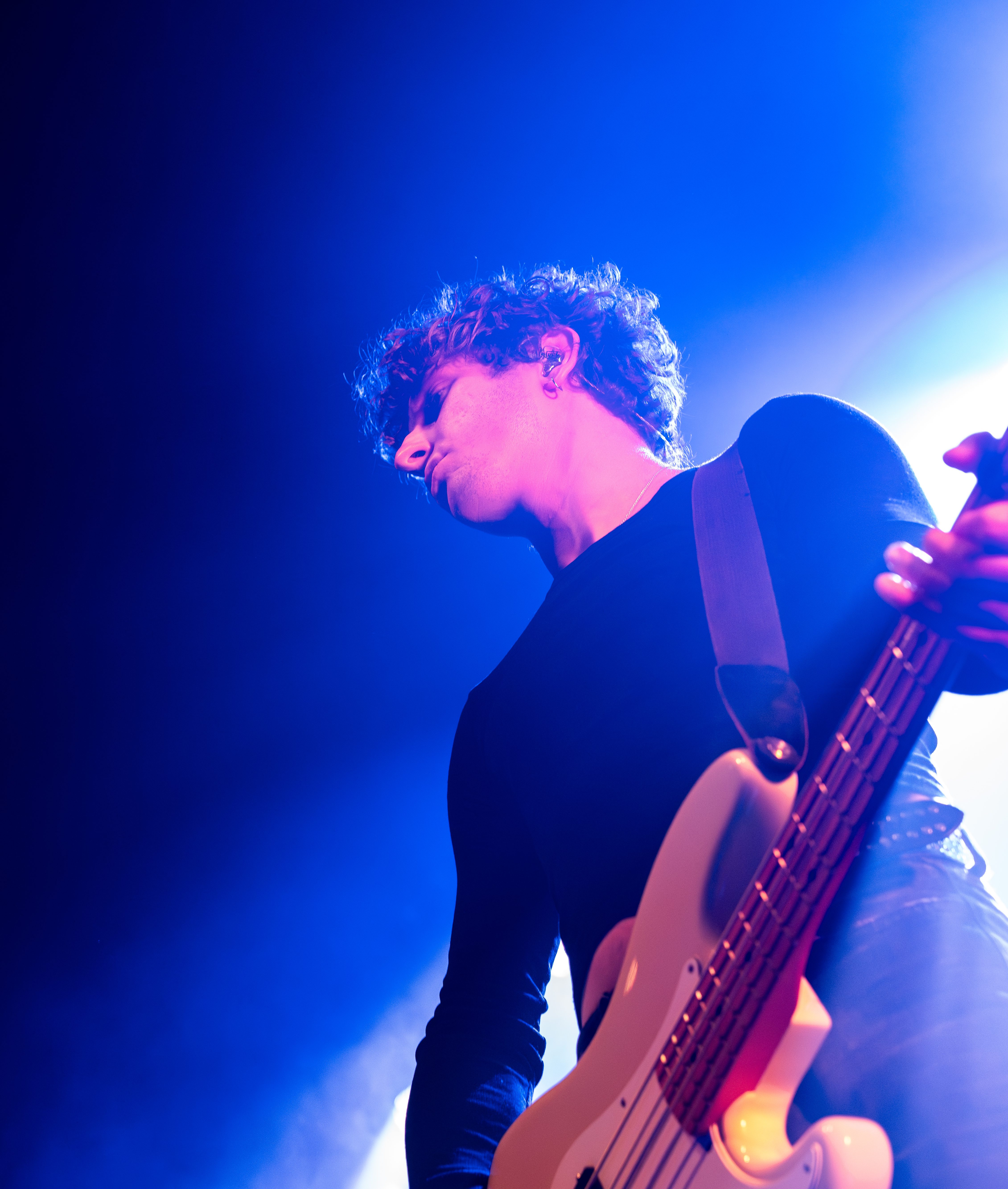 A musician playing the bass guitar on stage, illuminated by blue and purple lights.
