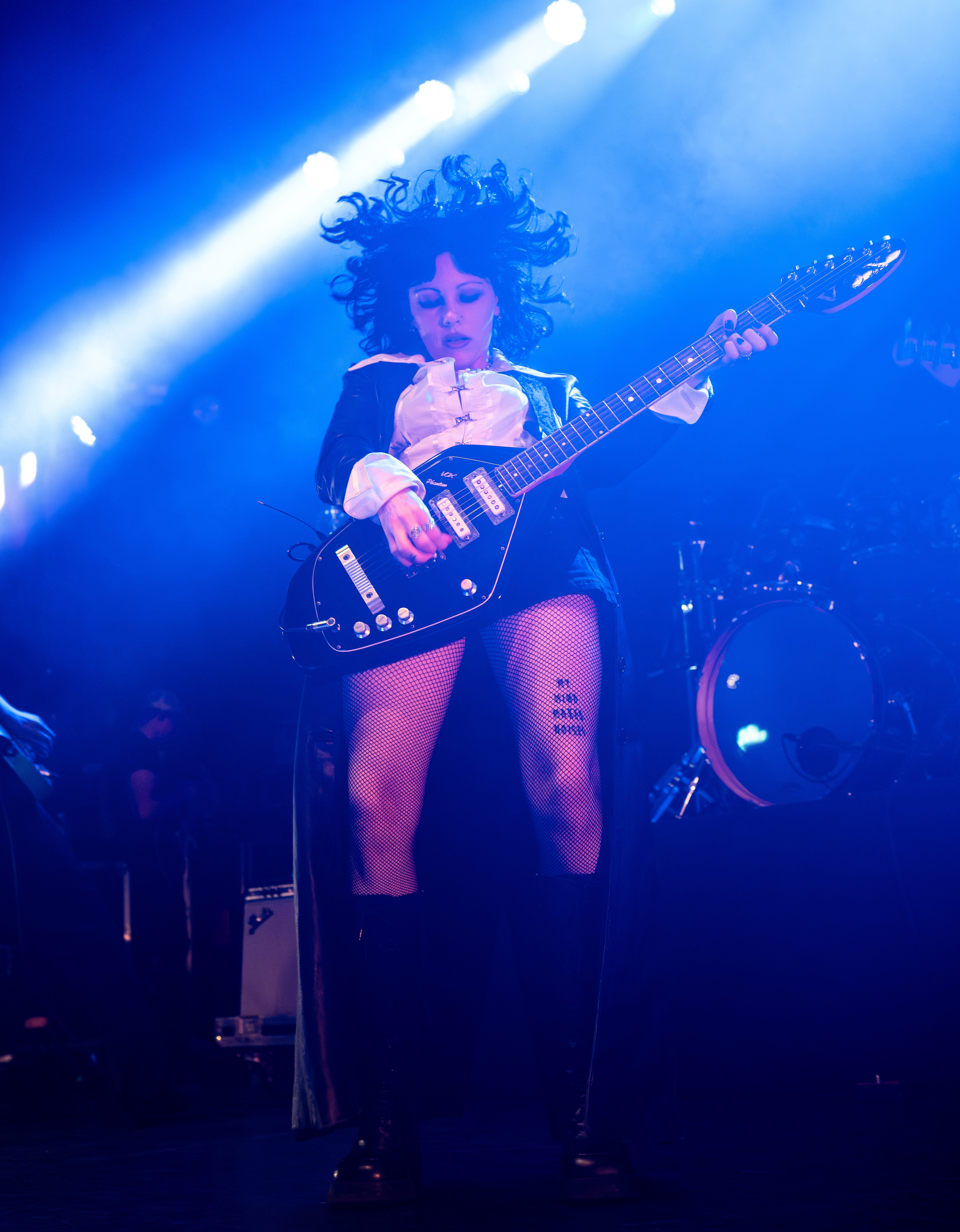 A musician performing on stage with a guitar, wearing a stylish black outfit and fishnet stockings, illuminated by blue stage lights.