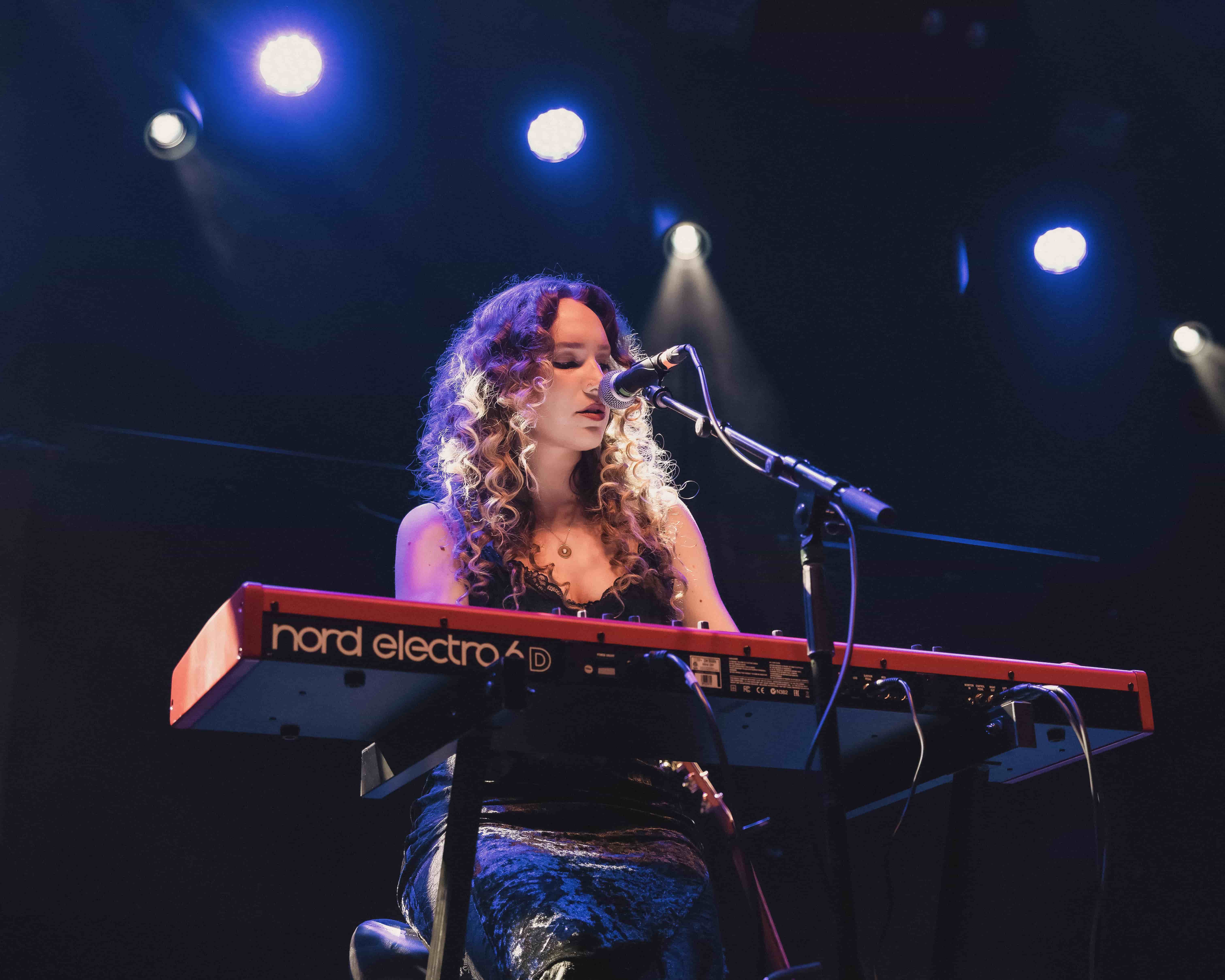 A female musician with curly hair performs on stage, singing into a microphone while playing a red keyboard. The background features bright stage lights.