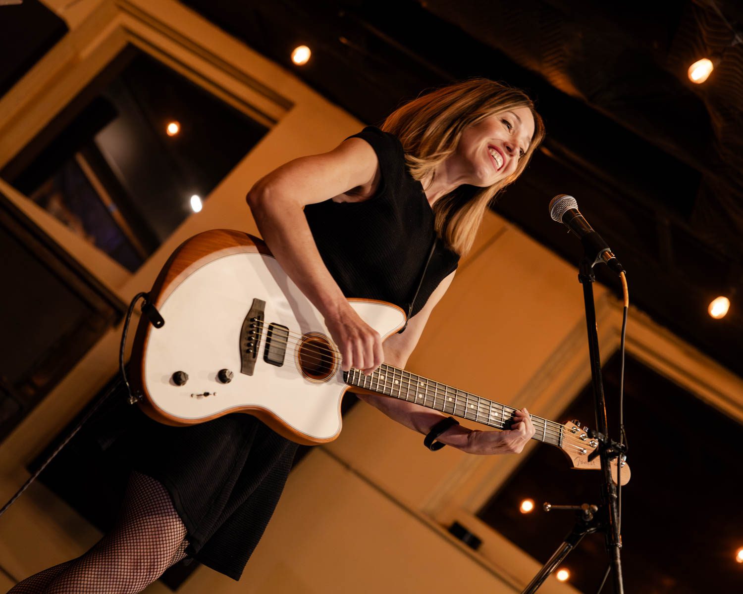 A smiling female musician playing an electric guitar on stage in a cozy venue with warm lighting.