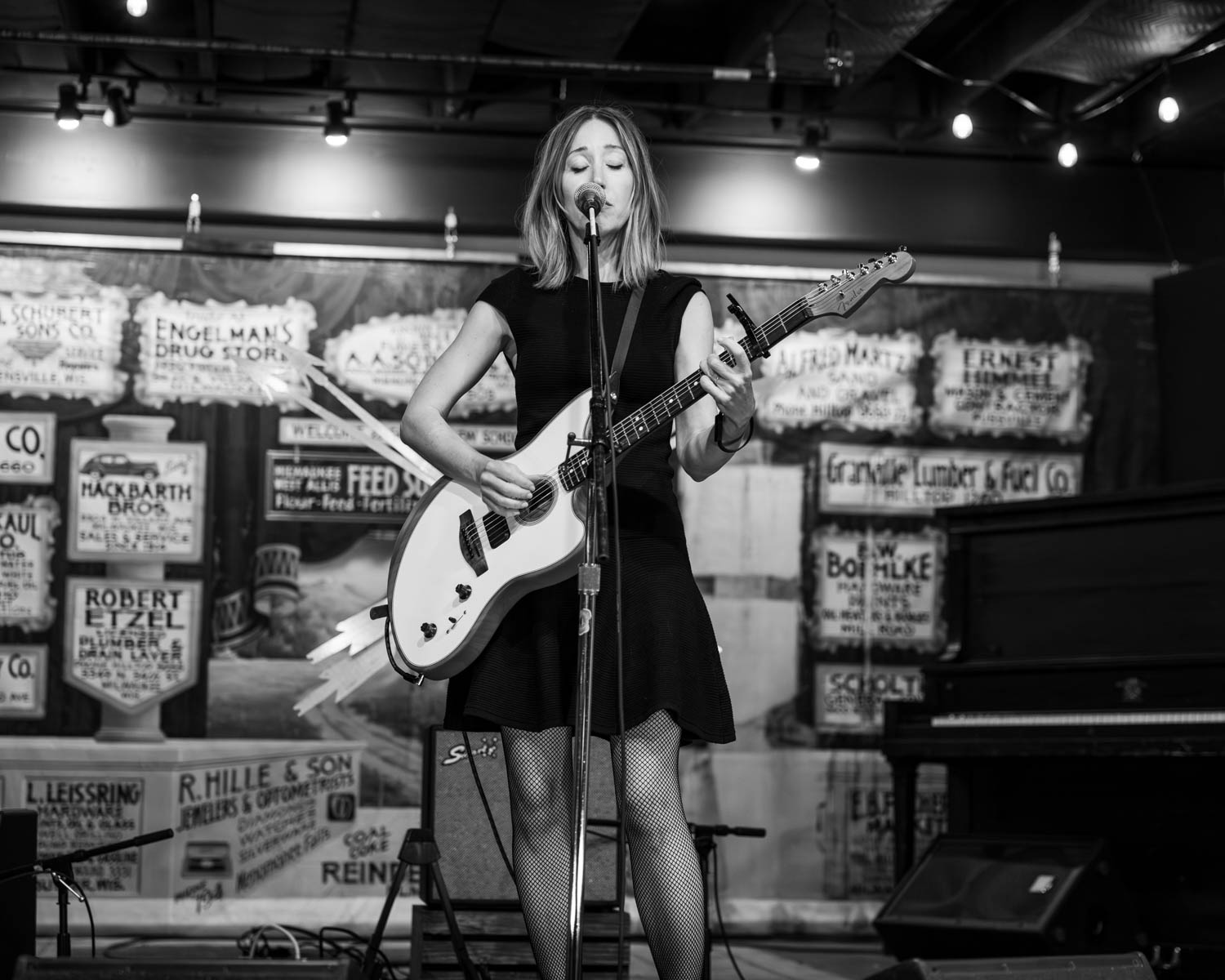 A solo female musician performing on stage, dressed in a black outfit and playing an electric guitar, while singing into a microphone. The background features vintage signs and a stage setup in a cozy, coffee shop environment.
