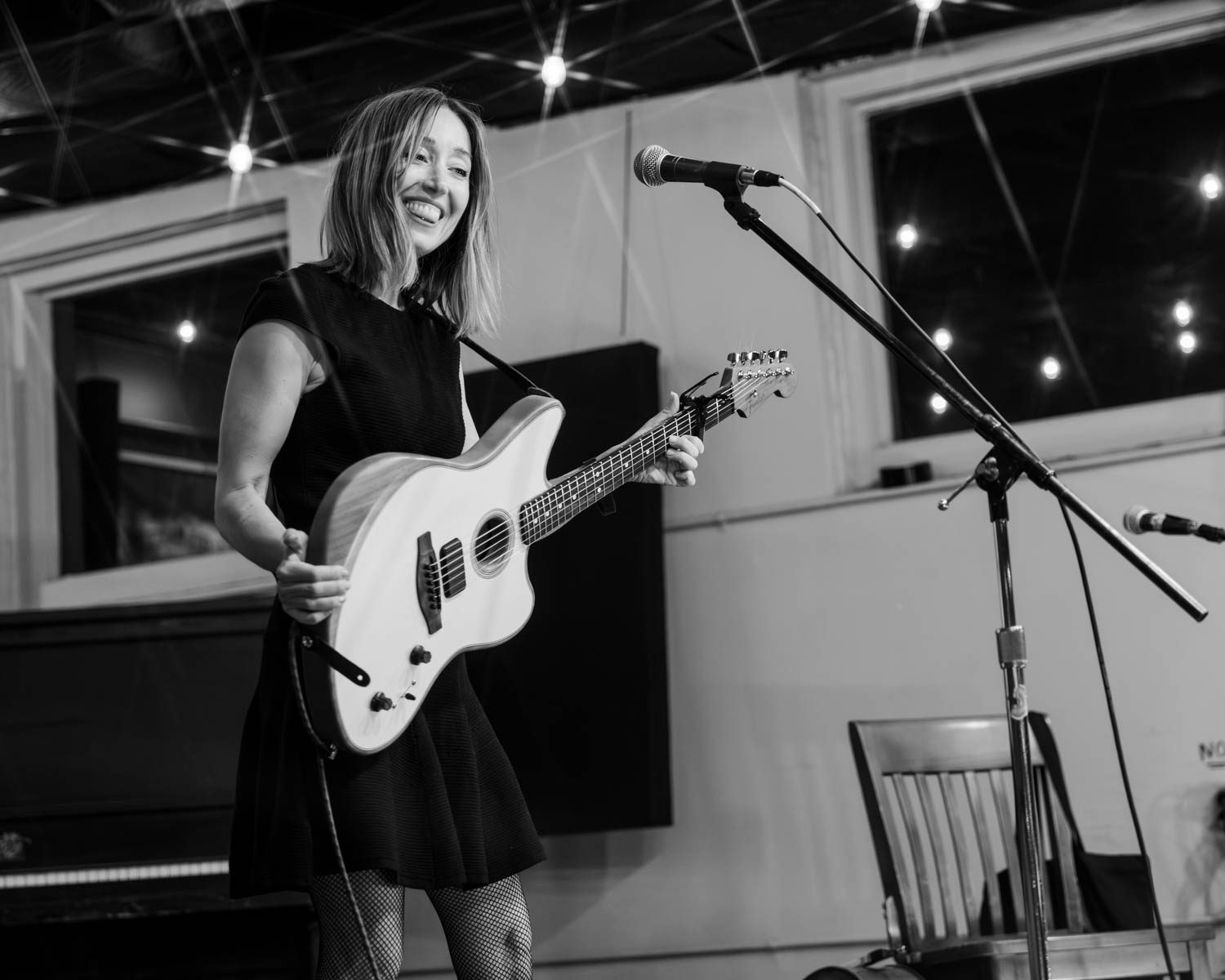 A smiling female musician holding a unique guitar while performing on stage in a cozy venue with decorative lighting.