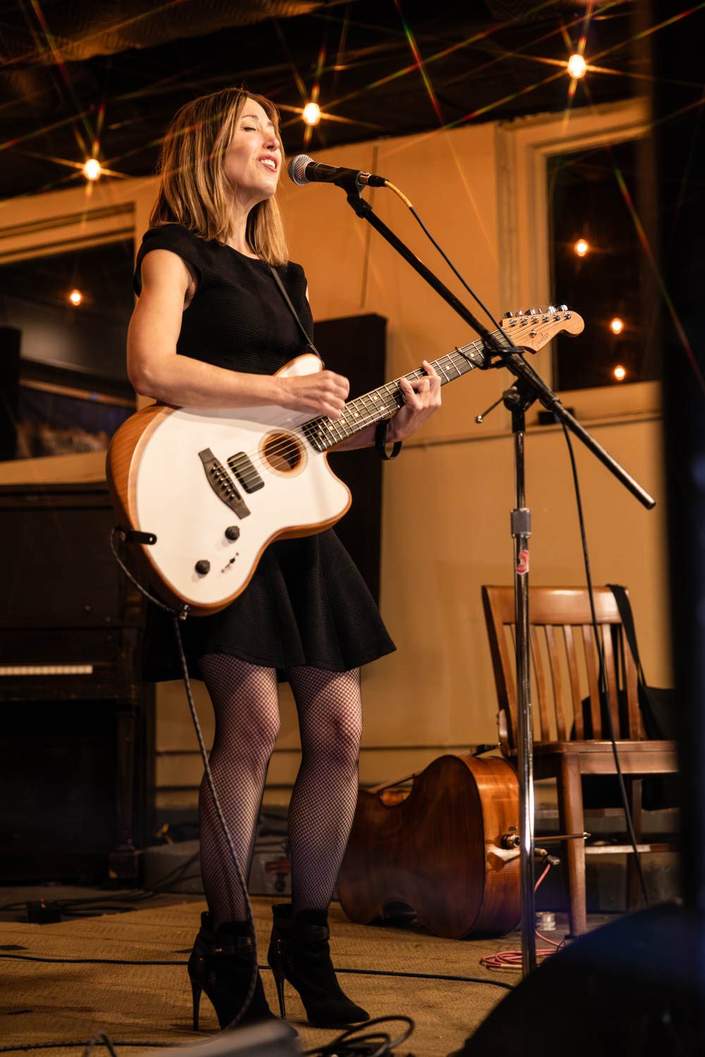 A woman performing on stage with a guitar, wearing a black dress and fishnet stockings, smiling while singing into a microphone.