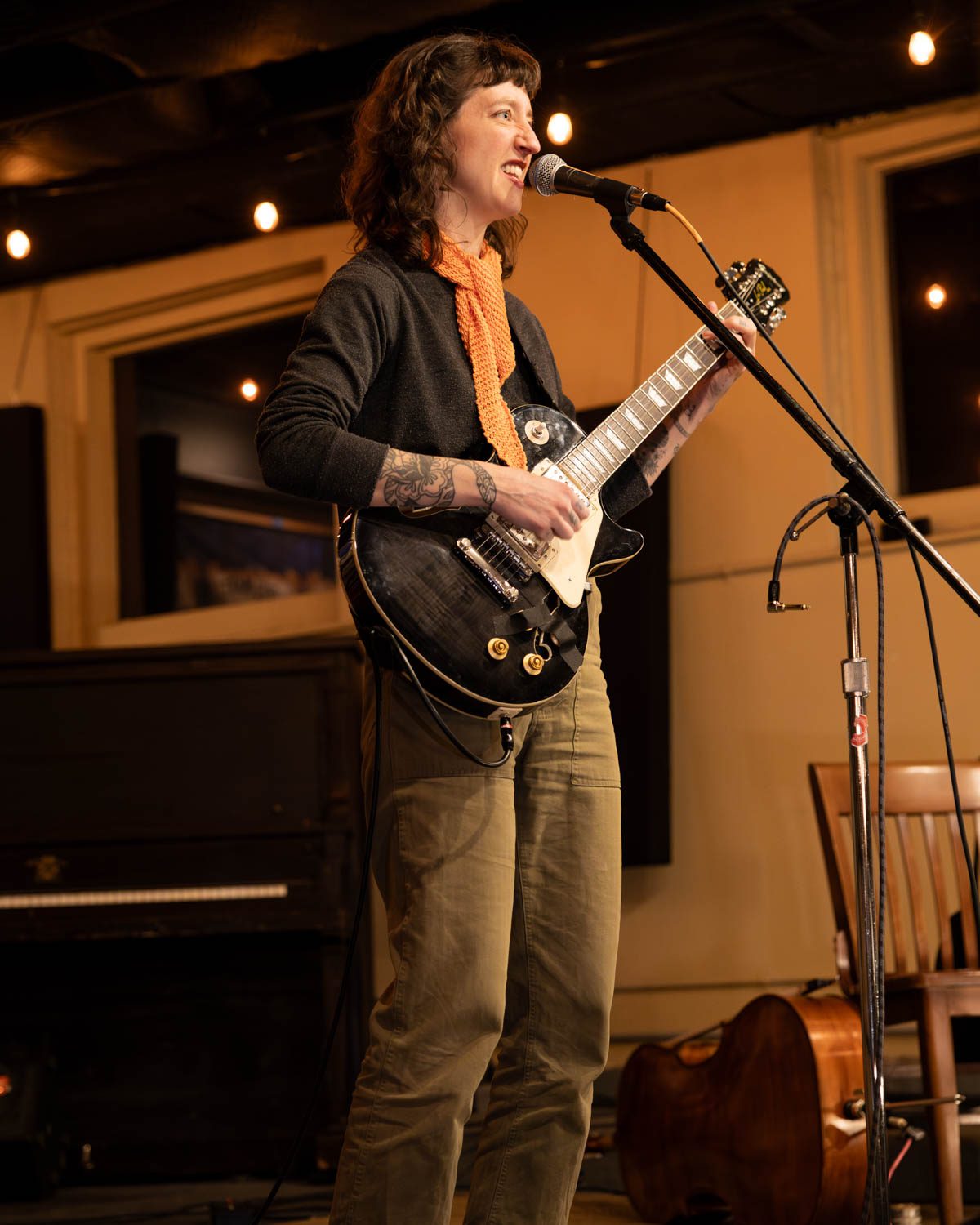 A female musician performing on stage with a guitar, smiling and engaging with the audience, in a cozy coffee shop setting.